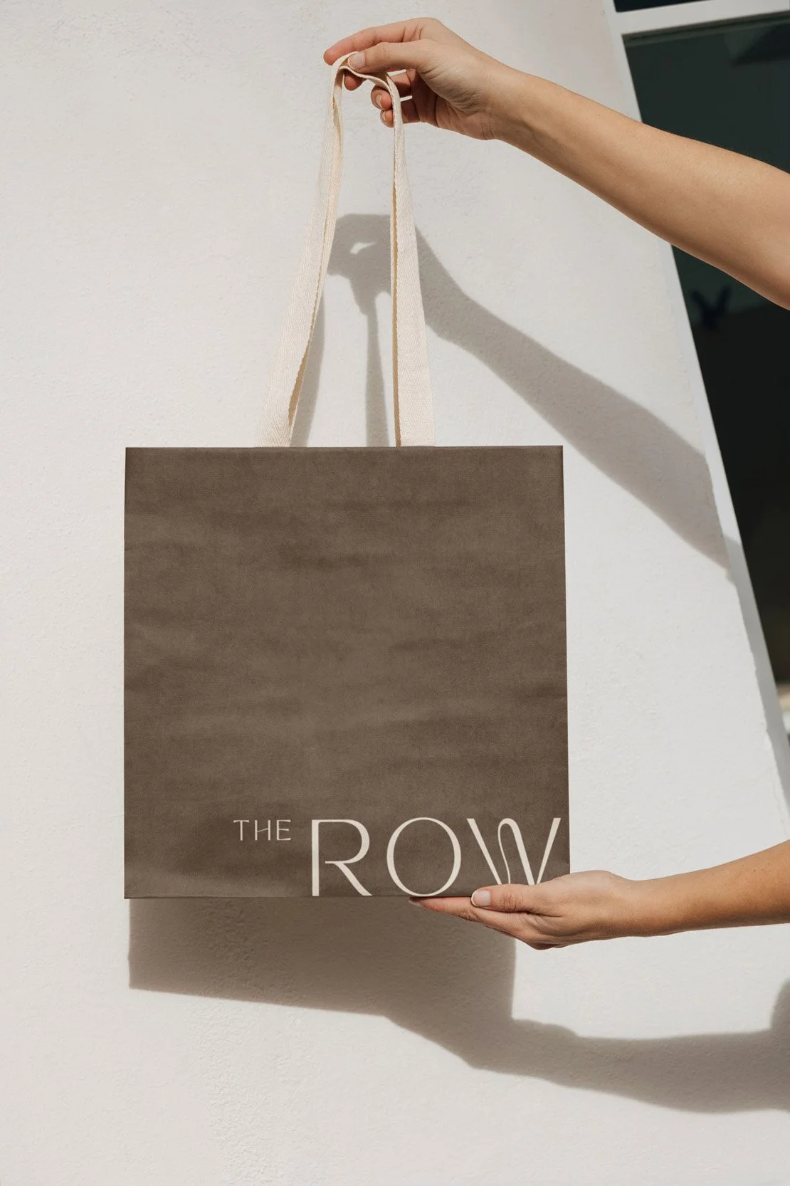 Person holding a shopping bag with the words 'THE ROW' printed on it, hanging on a white wall.