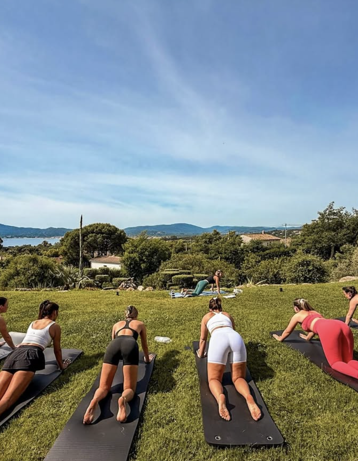Group of women participating in an outdoor yoga class on a grassy field, with a scenic view of hills, trees, and a lake in the background, under a clear blue sky.