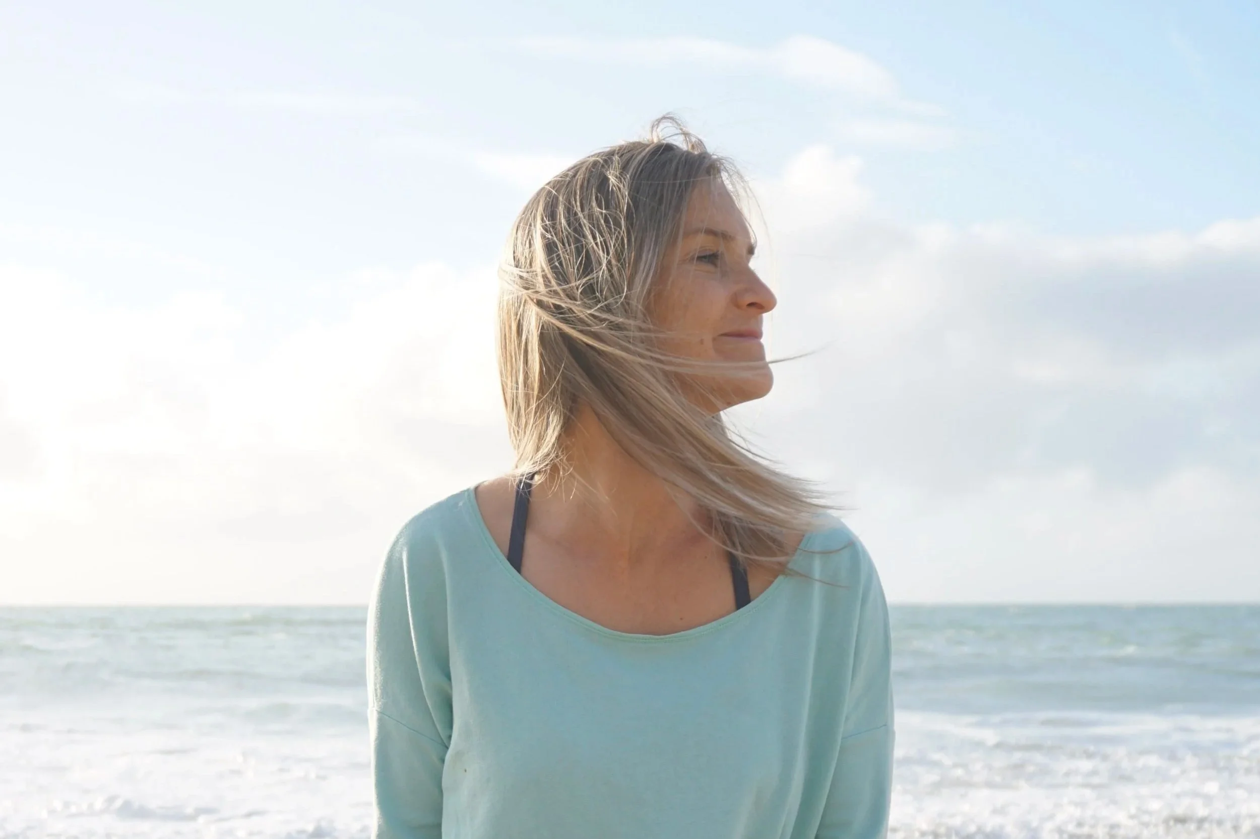 A woman with blonde hair standing on a beach with the ocean in the background, wearing a light blue top.