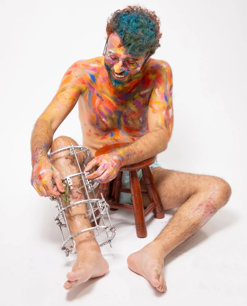 Homem com cabelo colorido, usando óculos, sentado em uma cadeira de madeira, brincando com uma perna presa por uma estrutura de metal, com tinta nas mãos e no corpo, em fundo branco.