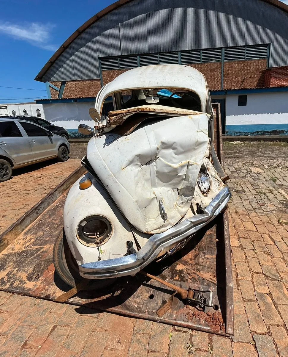 Carro antigo, branco, altamente danificado, com portas deformadas, sem rodas, transportado em uma plataforma de transporte em um local ao ar livre.