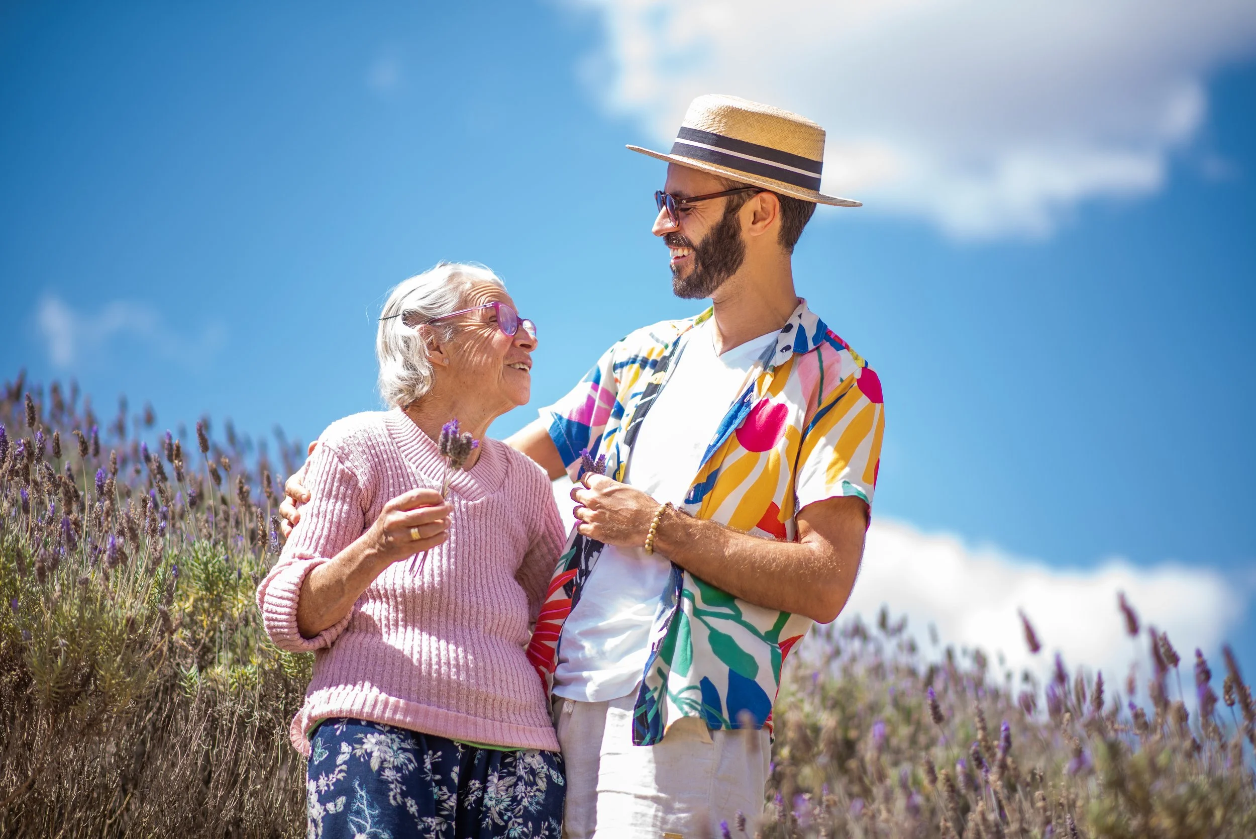 Jovem e idosa sorrindo, abraçando-se em um campo de lavanda sob céu azul