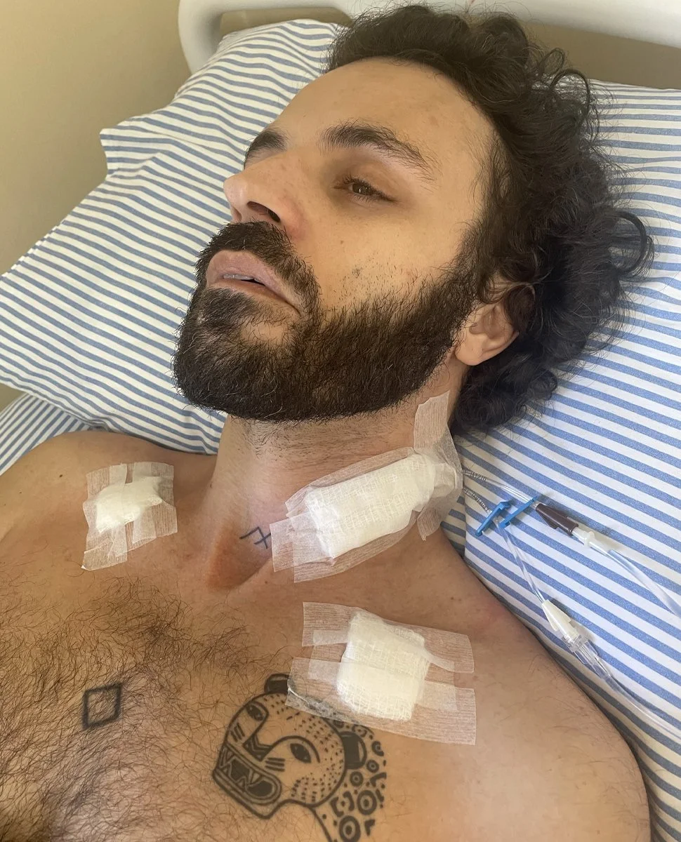 Paciente com barba e cabelo encarando para cima, deitado em um leito de hospital, com pontos em vários locais do pescoço e peito, conectado a tubos de intravenosa.