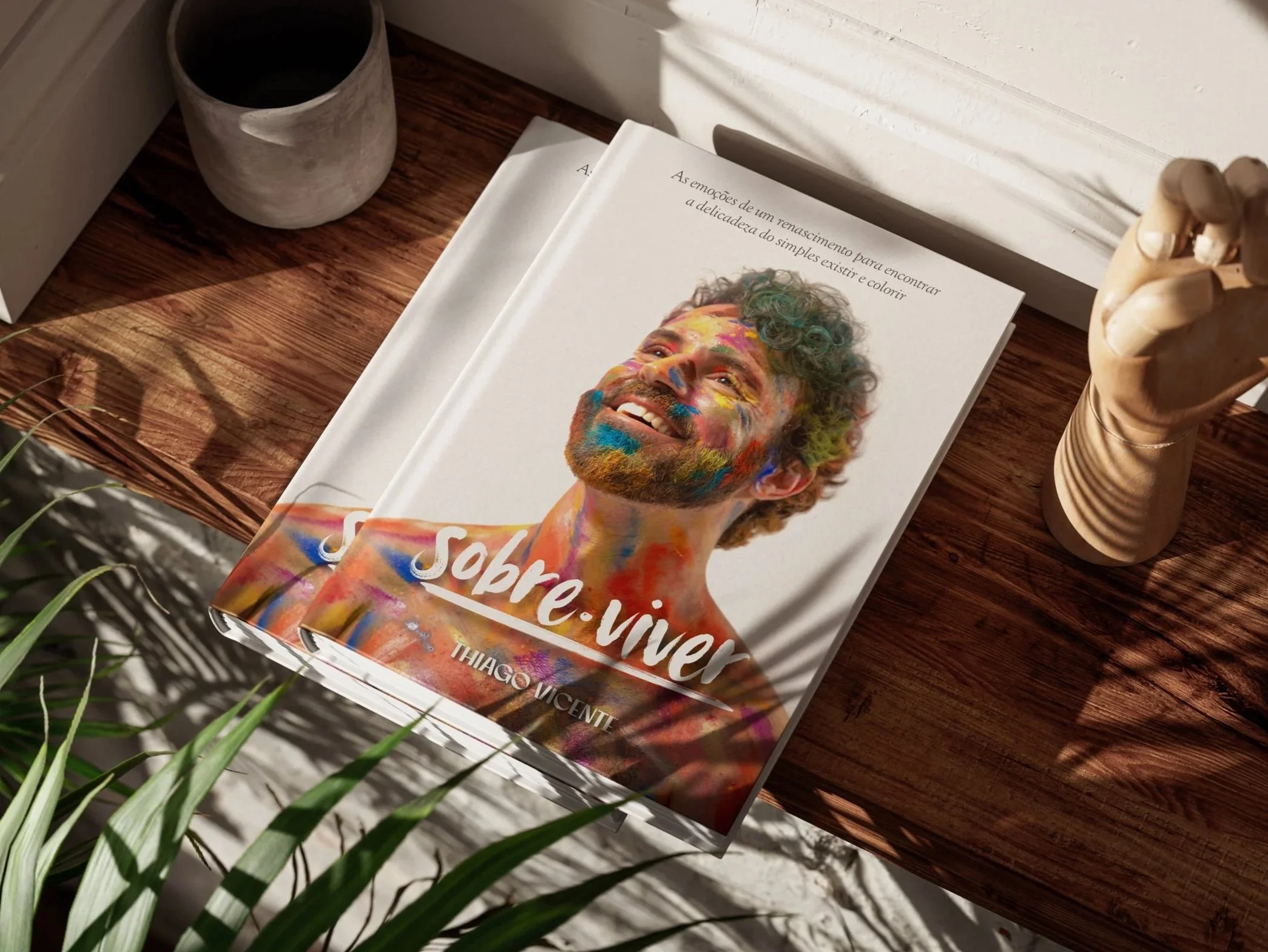 Livro com capa colorida apresentando um homem sorridente com maquiagem colorida no rosto, sobre uma mesa de madeira, acompanhada de vaso de cerâmica e planta ao redor.