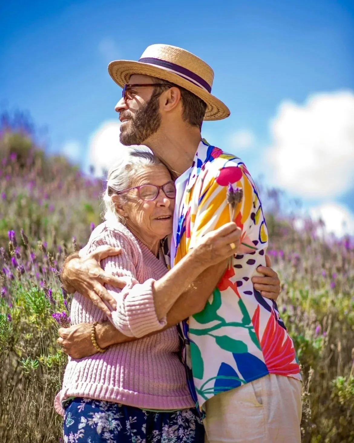 Um idoso e uma idosa se abraçam carinhosamente em um campo de flores, com o idoso usando chapéu de palha e roupas coloridas, e a idosa segurando uma flor. O céu está claro com poucas nuvens.