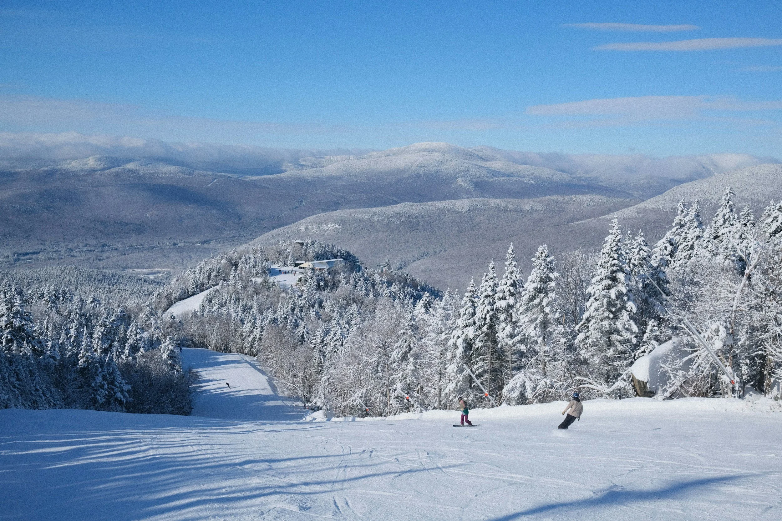 Snow-covered mountain landscape with trees and a few people skiing or snowboarding on a sunny day.