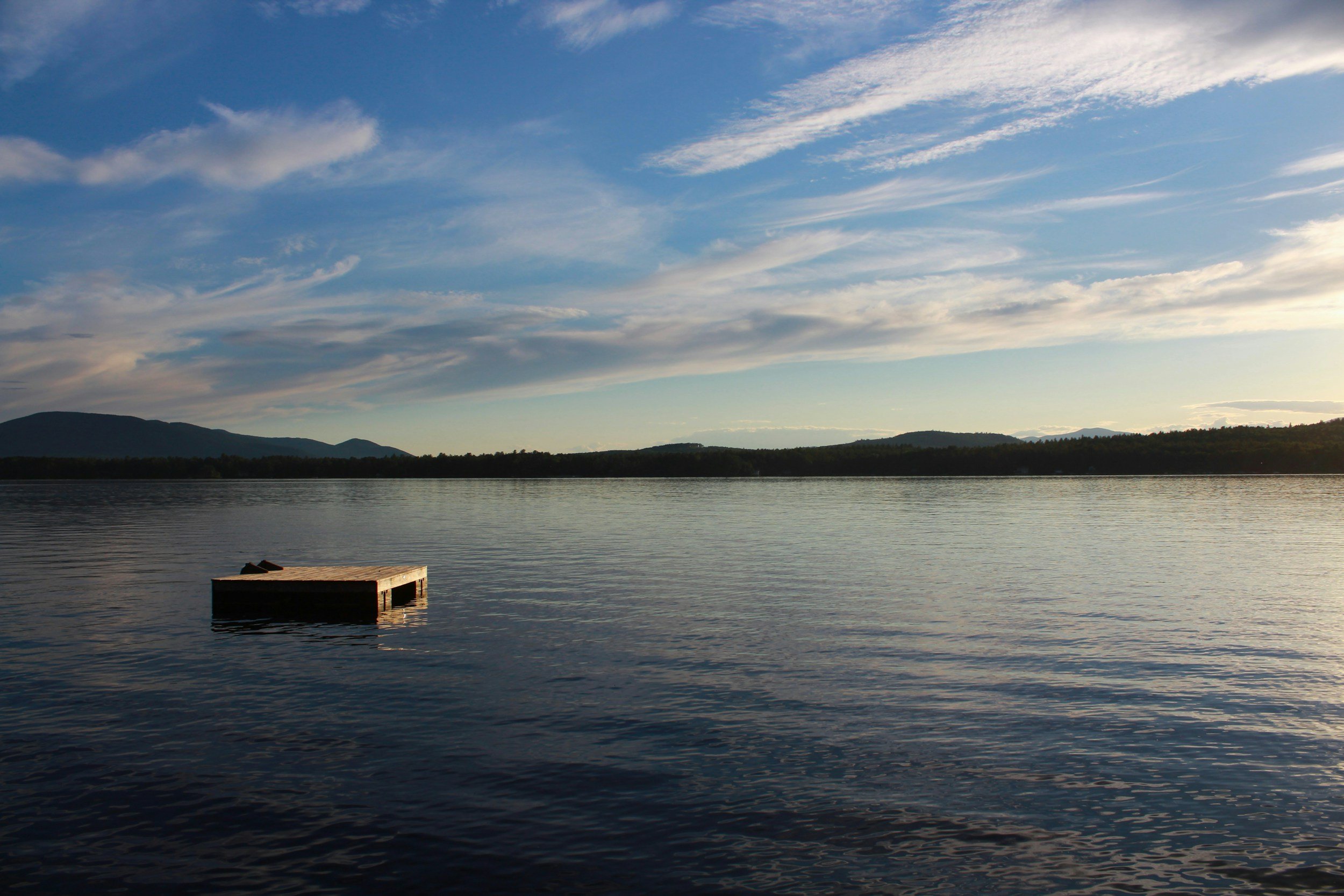 A calm lake or river during sunset with hills or mountains in the background and scattered clouds in the sky.
