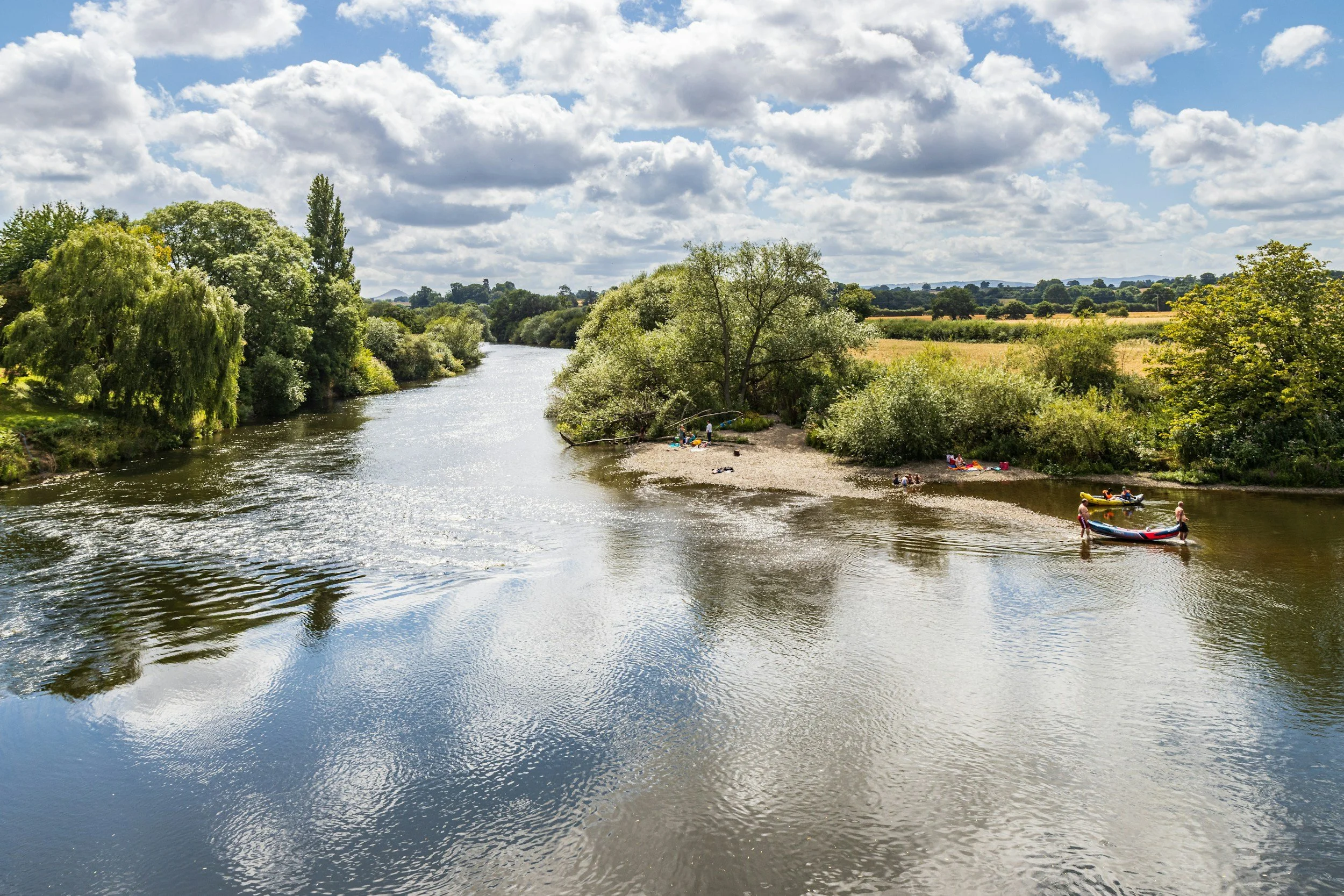 A peaceful river scene with trees on both sides and a partly cloudy sky. People are on the riverbank, some with kayaks and canoes preparing to paddle.