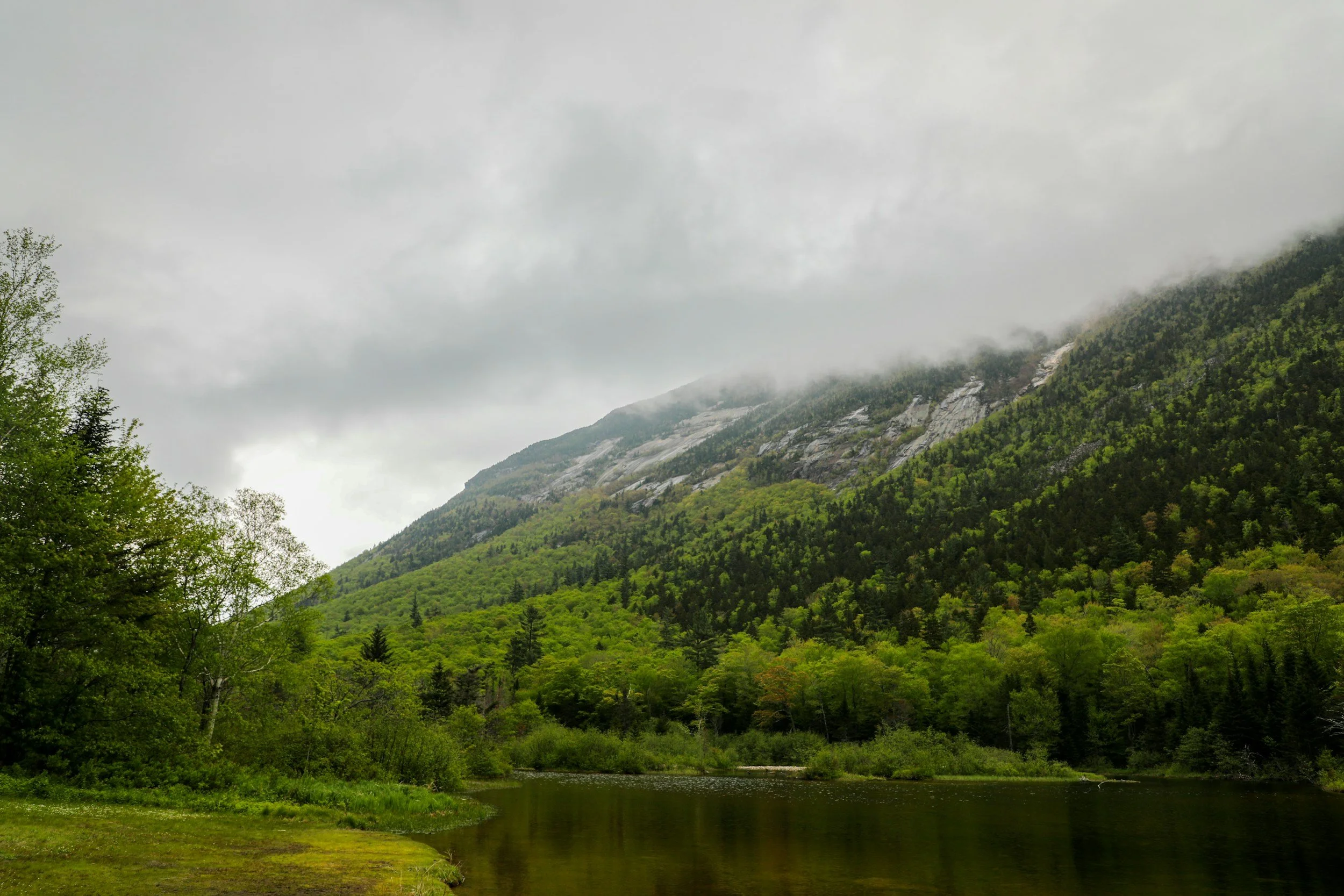 A scenic landscape featuring a river in the foreground, lush green forested hills, and a mountain partially covered in fog or low clouds in the background.