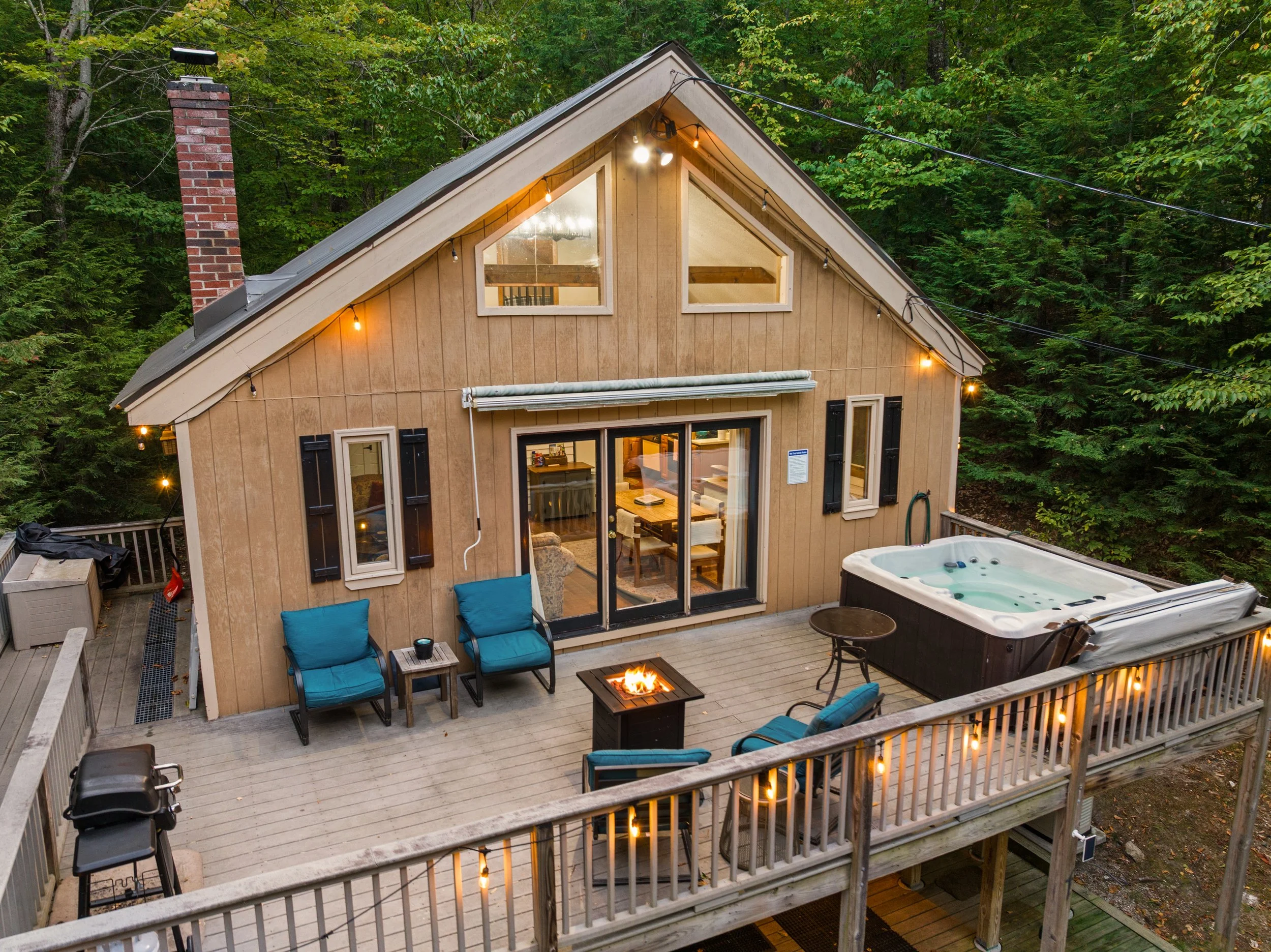A wooden deck surrounding a house with large glass doors, outdoor chairs, a fire pit, a hot tub, and string lights, with a dense forest background. Vacation Home in White mountains New Hampshire