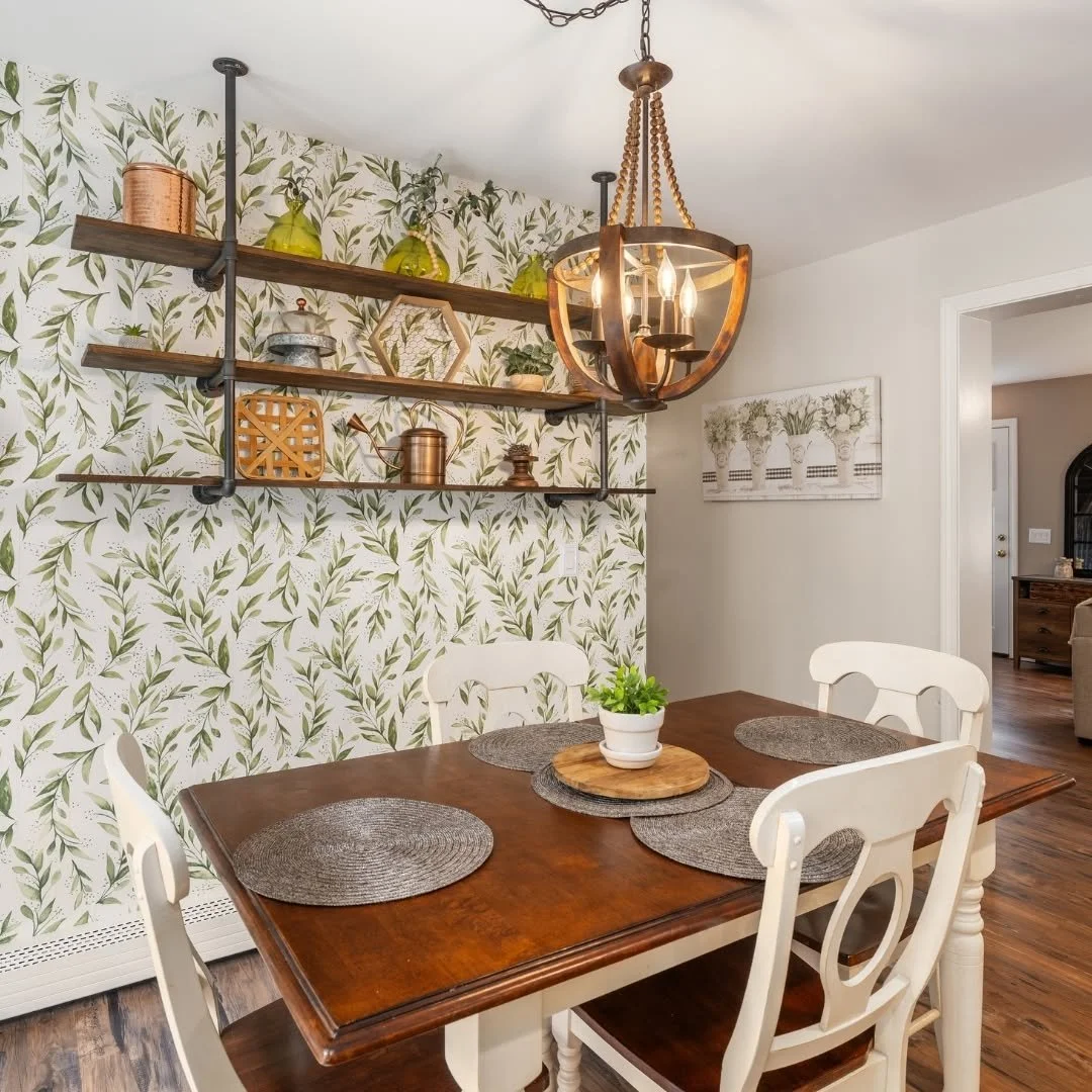Dining room with a wooden table, four white chairs, a plant centerpiece, patterned wallpaper with green leaves, open wooden shelves with decorative items, a chandelier, and a picture on the wall.