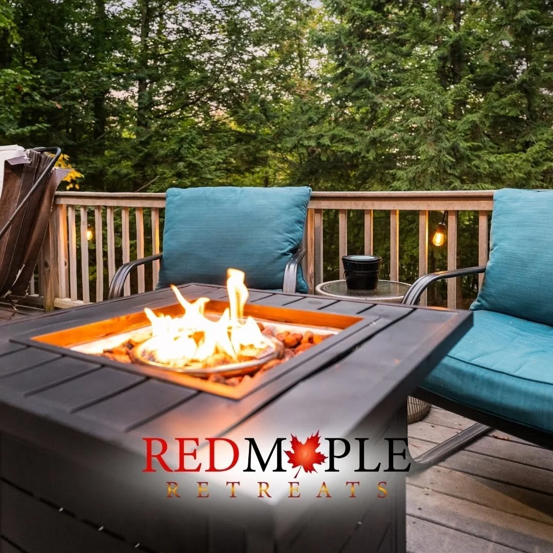 A cozy outdoor deck with a fire pit in the foreground, blue cushioned chairs, a small side table with a black container, string lights, and lush green trees in the background, with a logo for Red Maple Retreats.