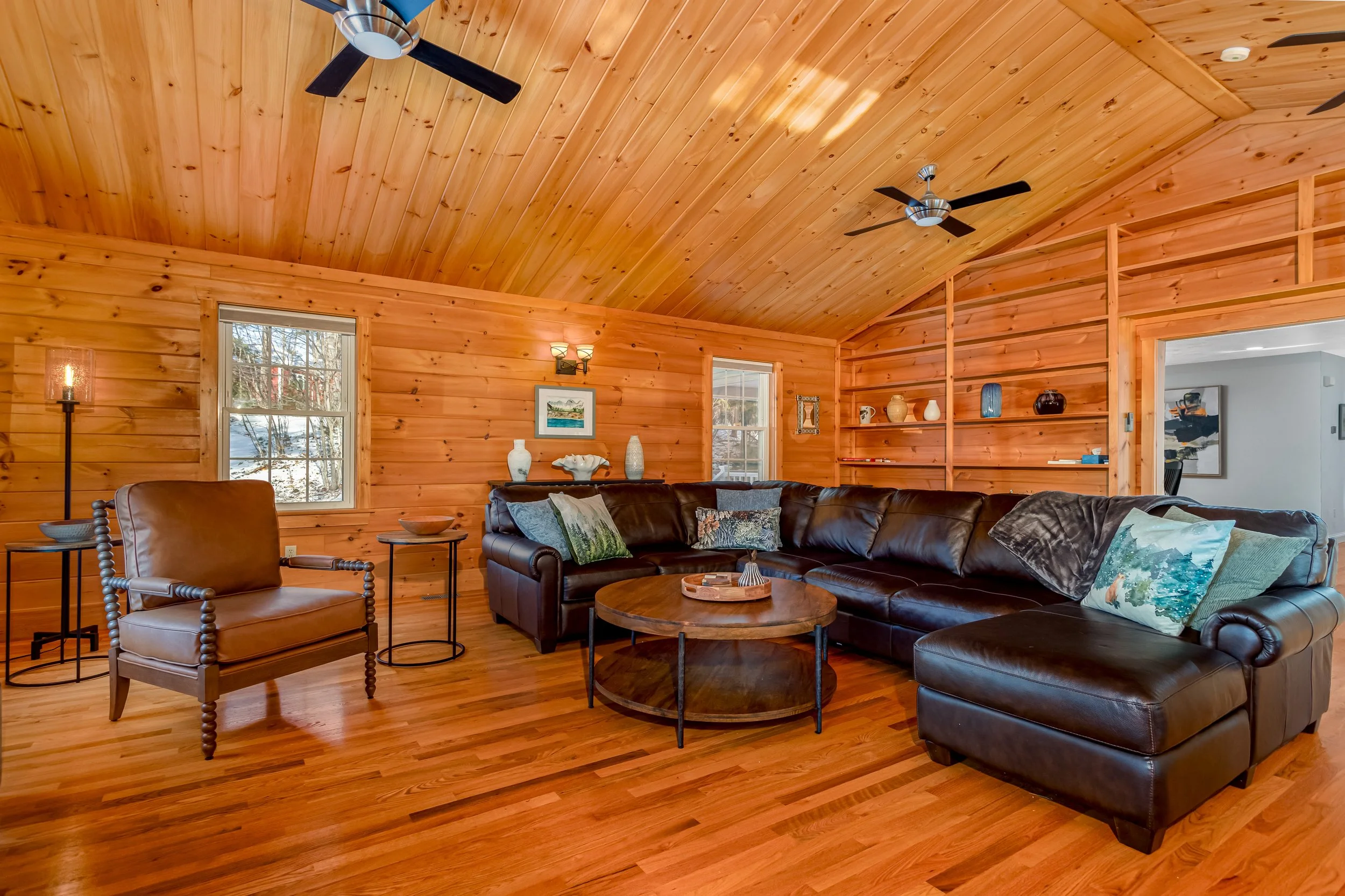 Living room with wooden ceiling and walls, a stone fireplace, large windows, a flat-screen TV, and various seating including a brown leather sofa and chairs.
