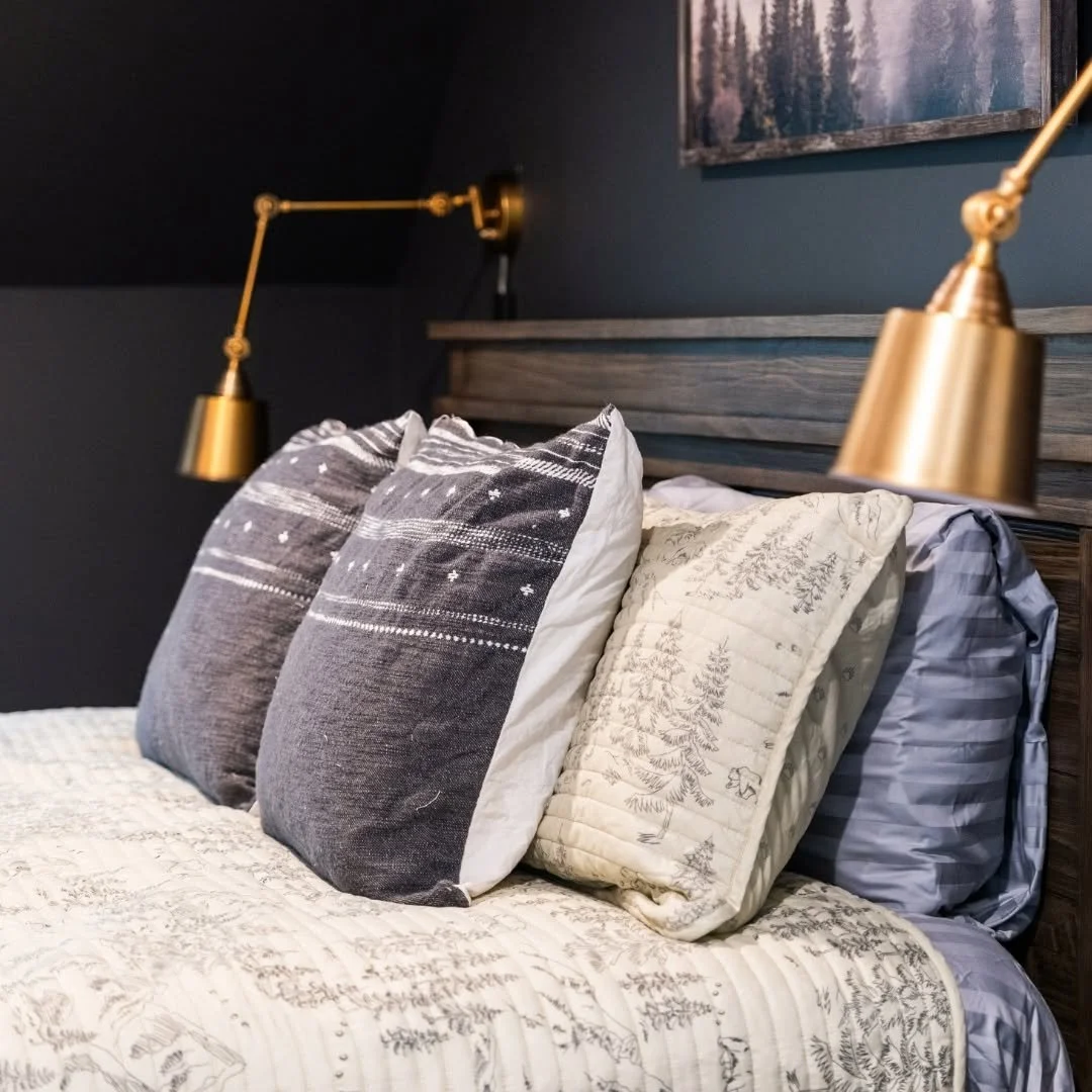 A bed with multiple pillows and a wooden headboard, with gold-colored reading lamps on either side mounted to the wall.