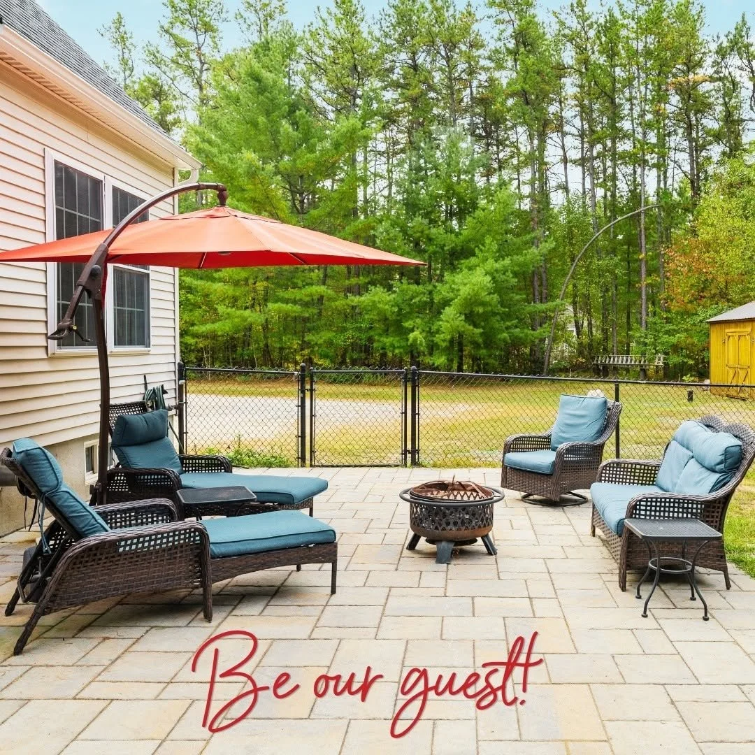 A patio with outdoor furniture including a lounge chair, four armchairs, and a small table, surrounded by a black fence with a forested area in the background. There is a large orange patio umbrella providing shade, and the text "Be our guest!" in red cursive at the bottom of the image.