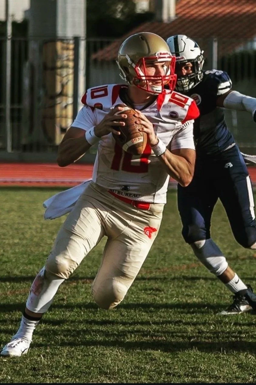 A football player in a white and red uniform holding a football while running, with another player in a black uniform attempting to tackle him during a game on a grassy field.
