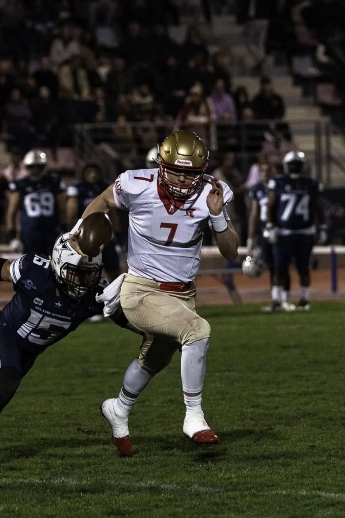 A football player in a white and gold uniform, number 7, running on a football field with a football in his right hand, while an opposing player tries to tackle him from behind. The scene is set at night with spectators in the background.