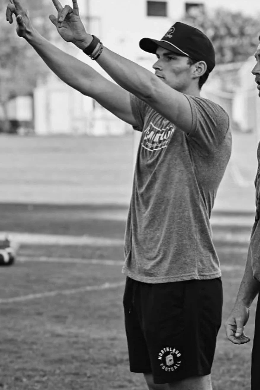 A young man in a baseball cap and t-shirt is standing on a sports field with his arm raised, making a hand gesture, while another person stands nearby.
