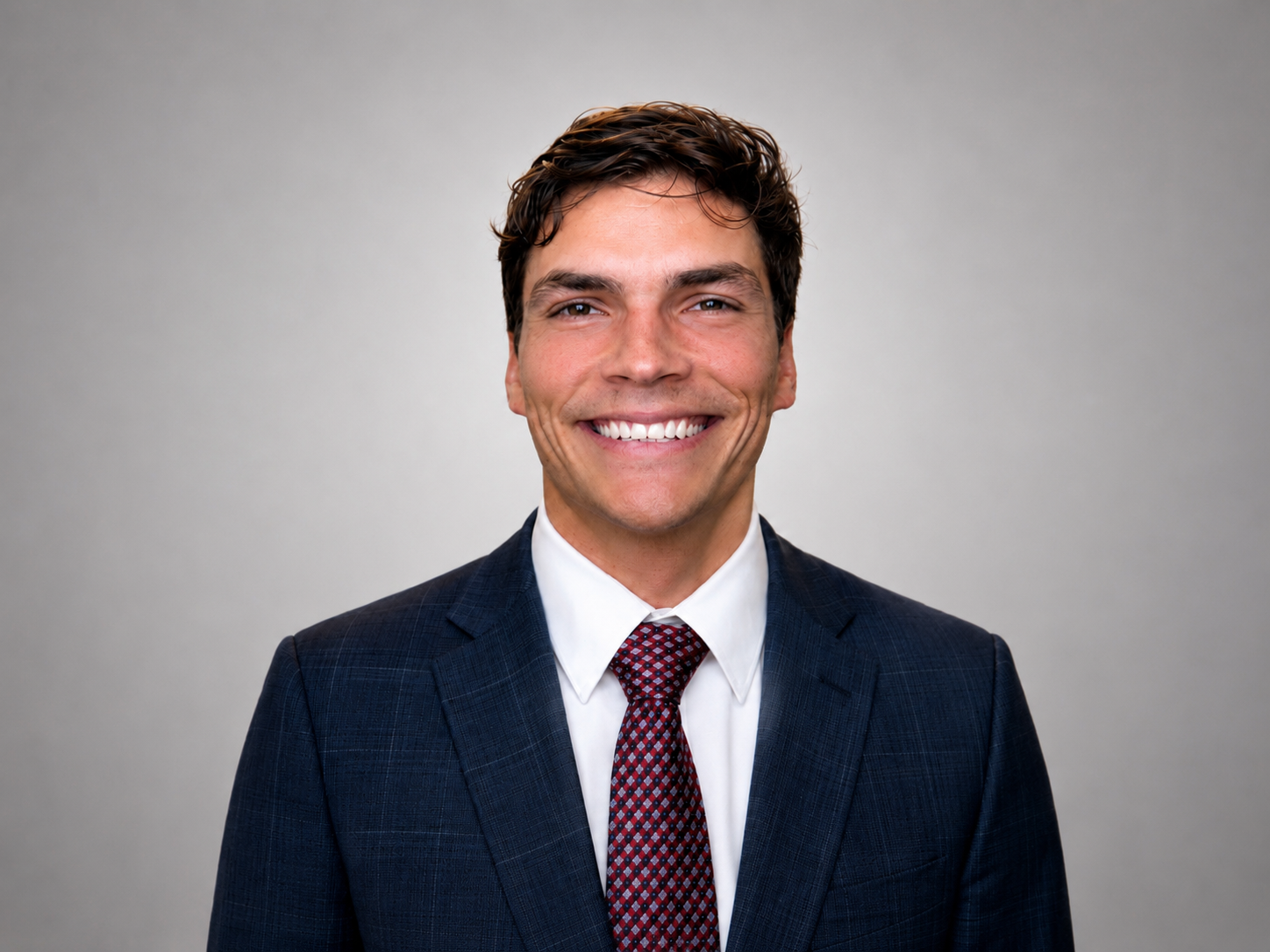 Portrait of a young man in a business suit smiling, with a plain gray background.