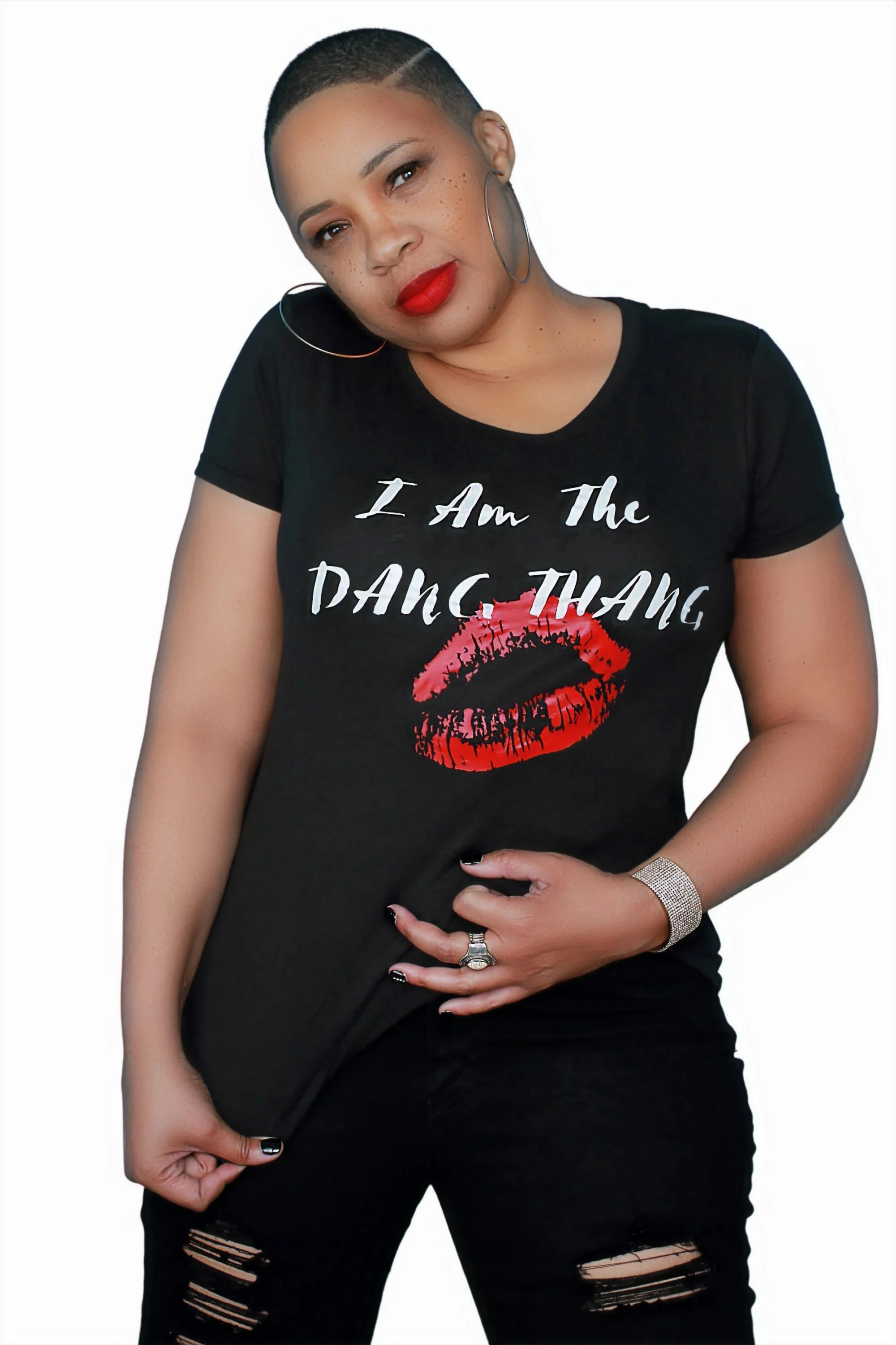 A woman with short hair, freckles, and red lipstick wearing a black T-shirt with a red lipstick kiss print and the text "I Am The DANG THANG." She has hoop earrings, a bracelet, rings, and is standing against a white background.