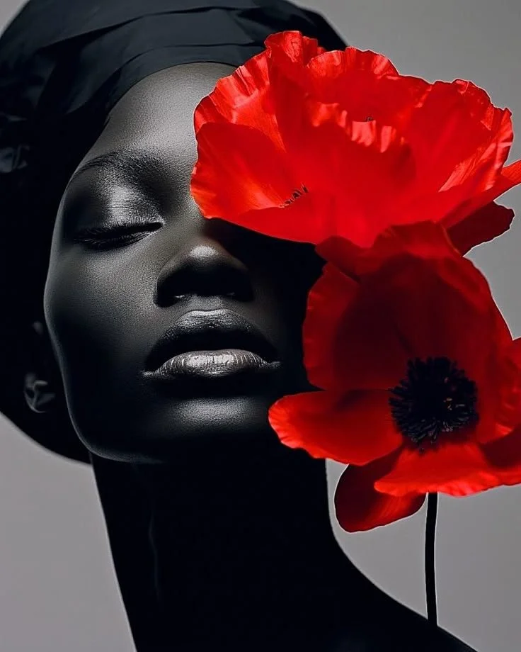 A black woman with eyes closed, wearing a head wrap, holding two bright red poppy flowers in front of her face.