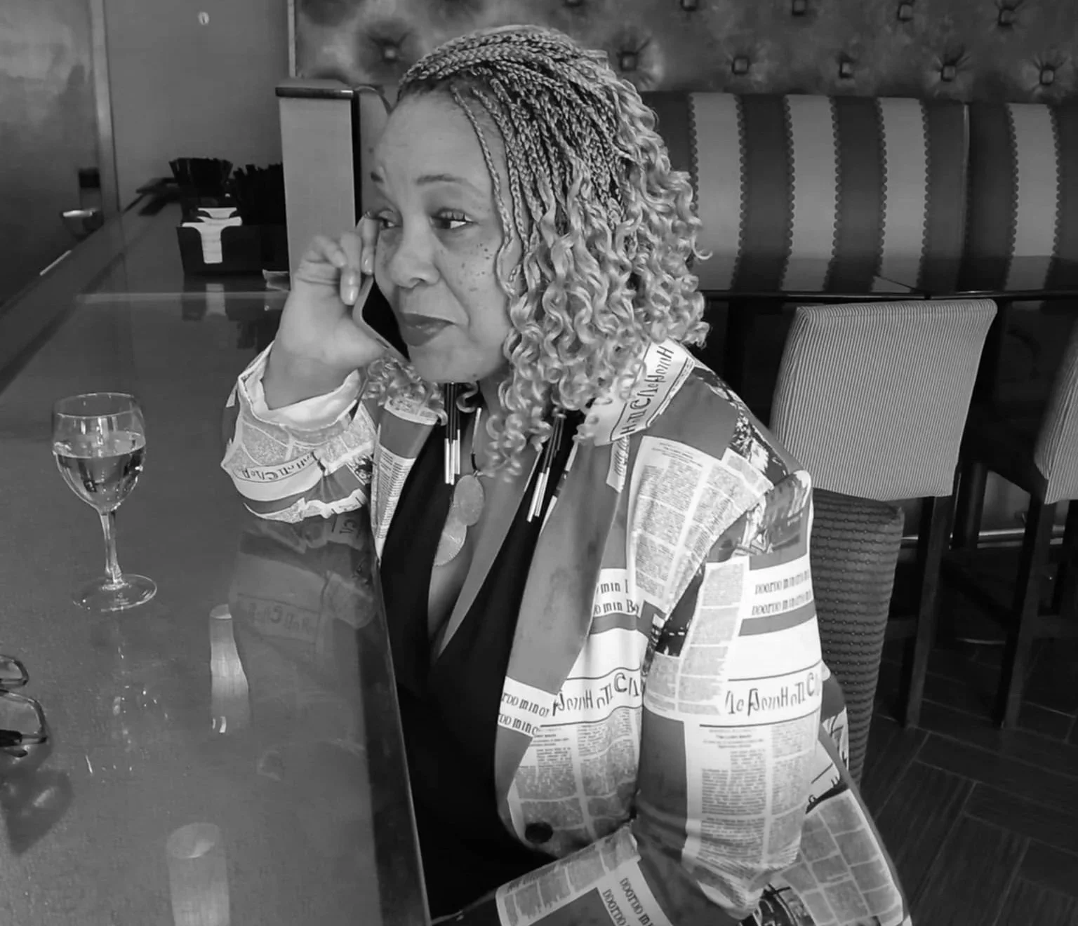 A woman with blonde braids, wearing a newspaper-patterned jacket, is sitting at a restaurant table talking on her phone.