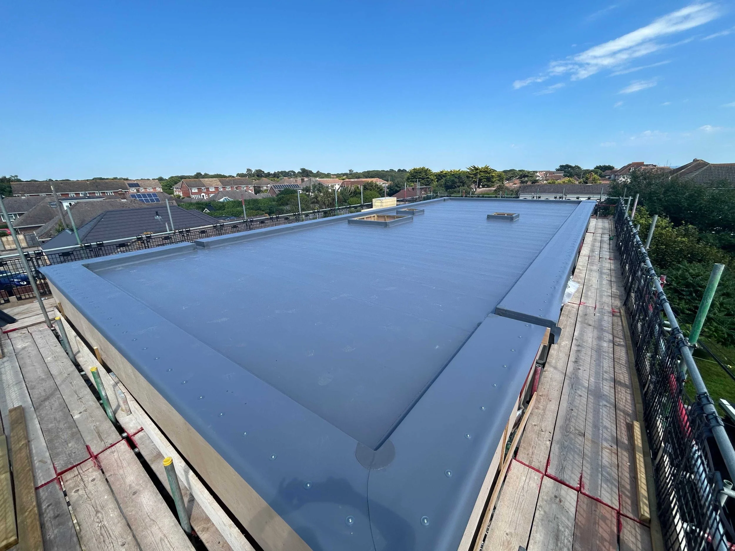 The photo shows a flat rooftop with a watertight membrane, surrounded by scaffolding and safety barriers, with a suburban neighborhood and blue sky in the background.