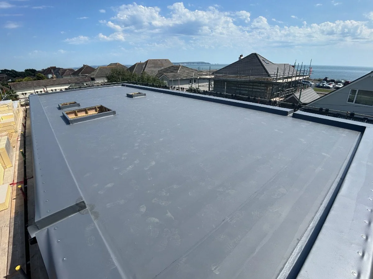 Flat metal roof with skylights under a blue sky, surrounding residential houses, some under construction, near a body of water.