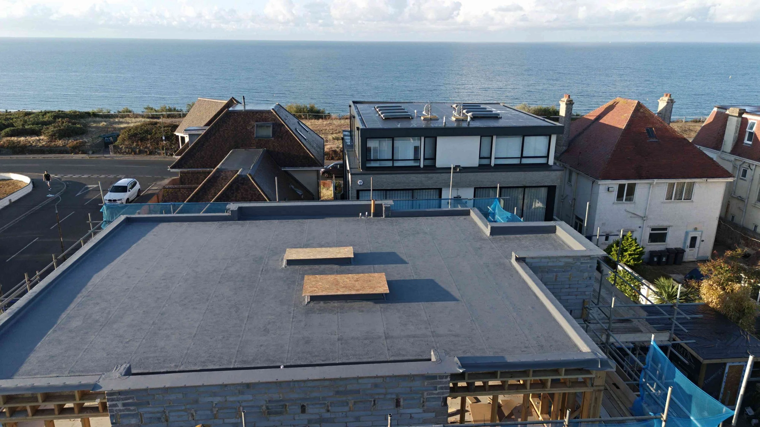View of residential buildings near the coast with a large flat rooftop in the foreground, some of which are under construction, and the ocean in the background.