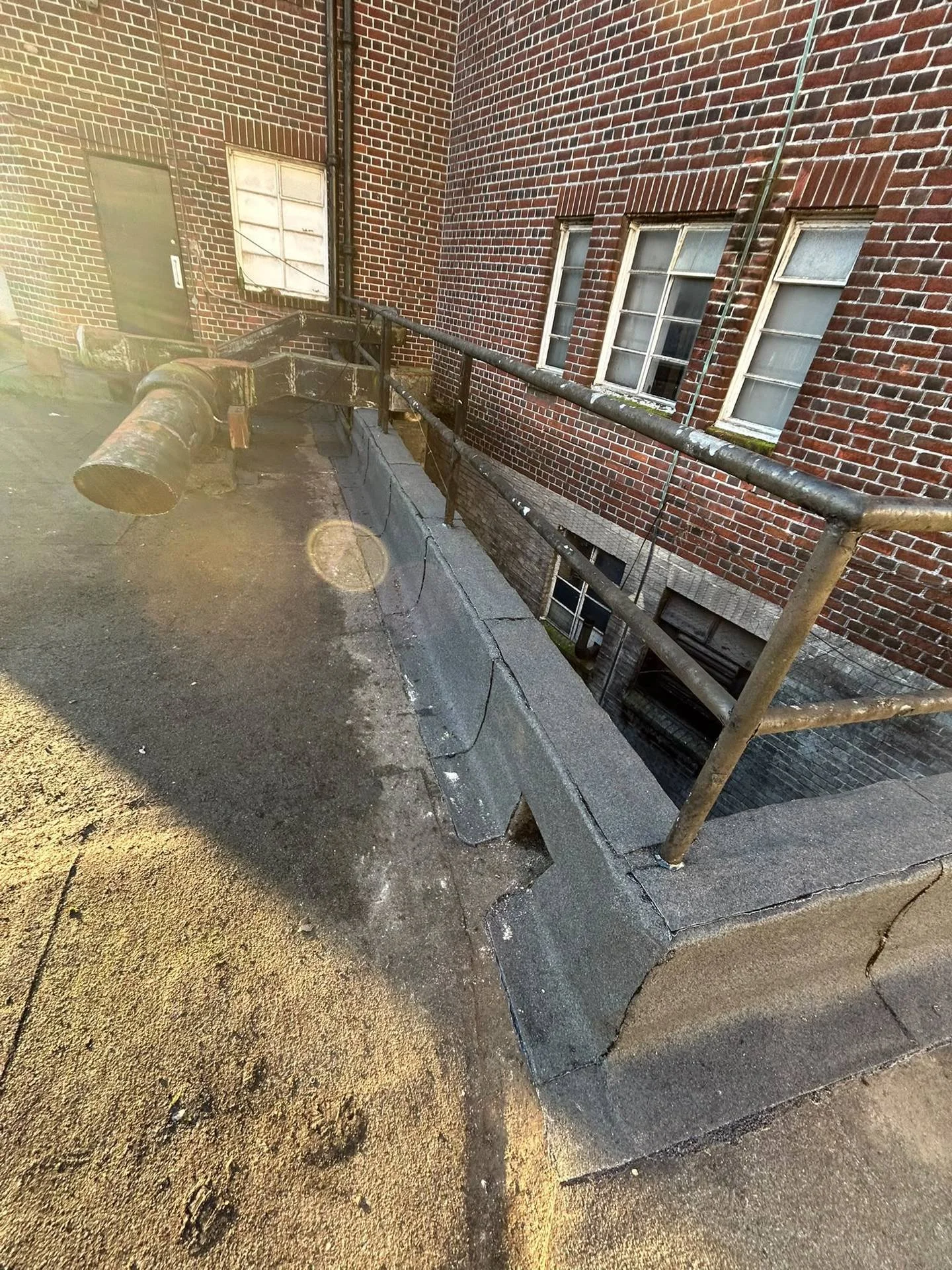 Closed sidewalk with damaged and missing concrete, rusty metal railing, brick building wall with clouds and three small windows, one with moss, and a metal pipe running vertically along the wall.