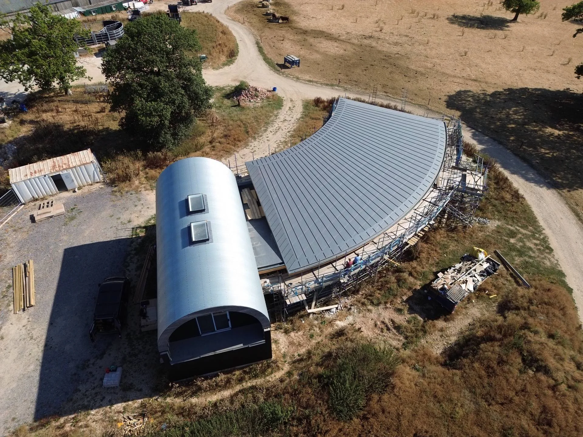 An aerial view of a construction site with a partially built structure featuring a curved, metallic roof and scaffolding. There are trees, a dirt road, and construction materials scattered around.