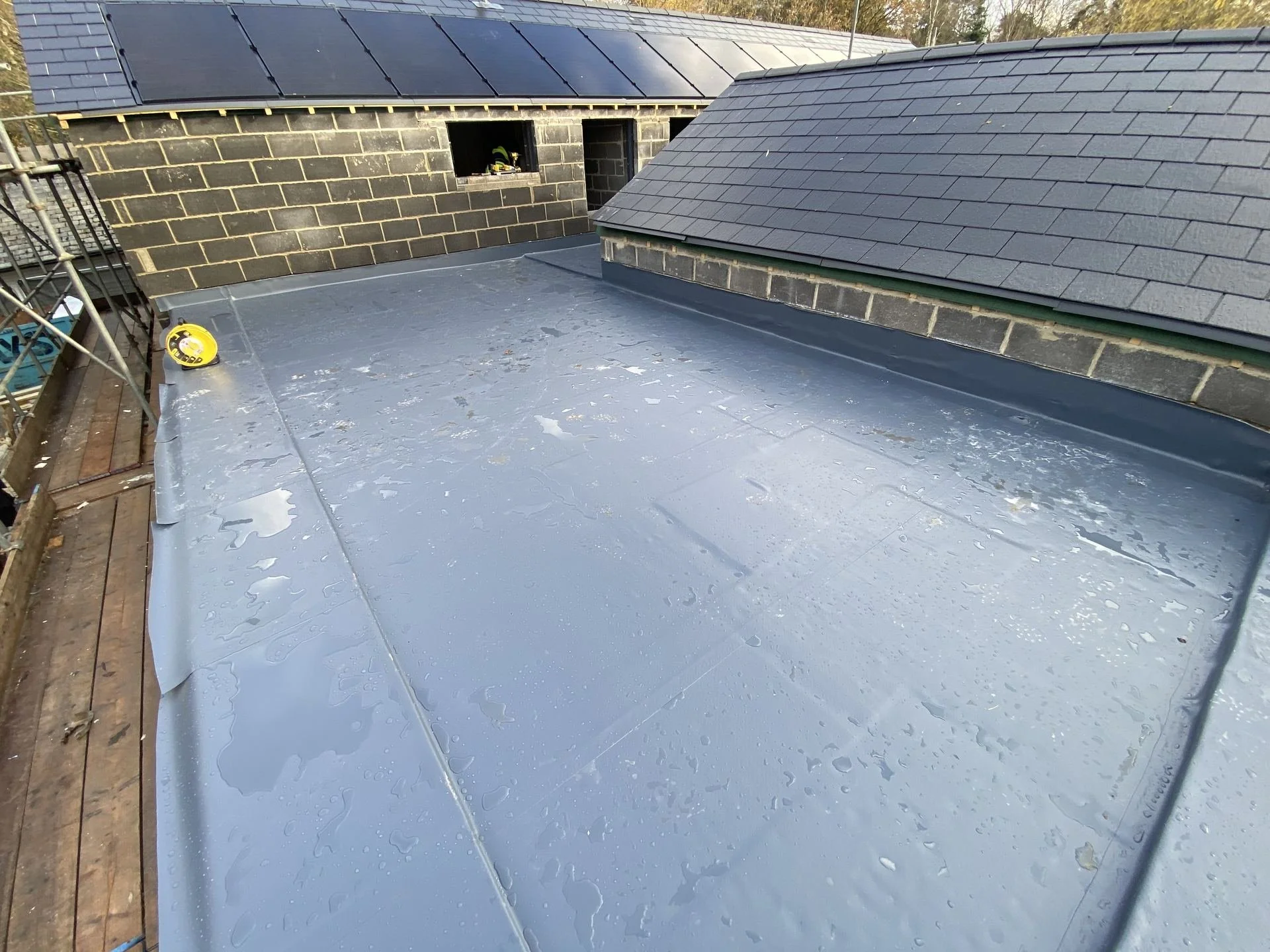 Rooftop with a black shingled hip roof, flat roofing material, and scaffolding on the side. There is a yellow tape measure on the flat surface.
