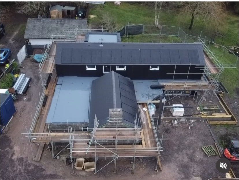 Aerial view of a house under construction with scaffolding surrounding it, a partially finished roof, and construction materials around the site.