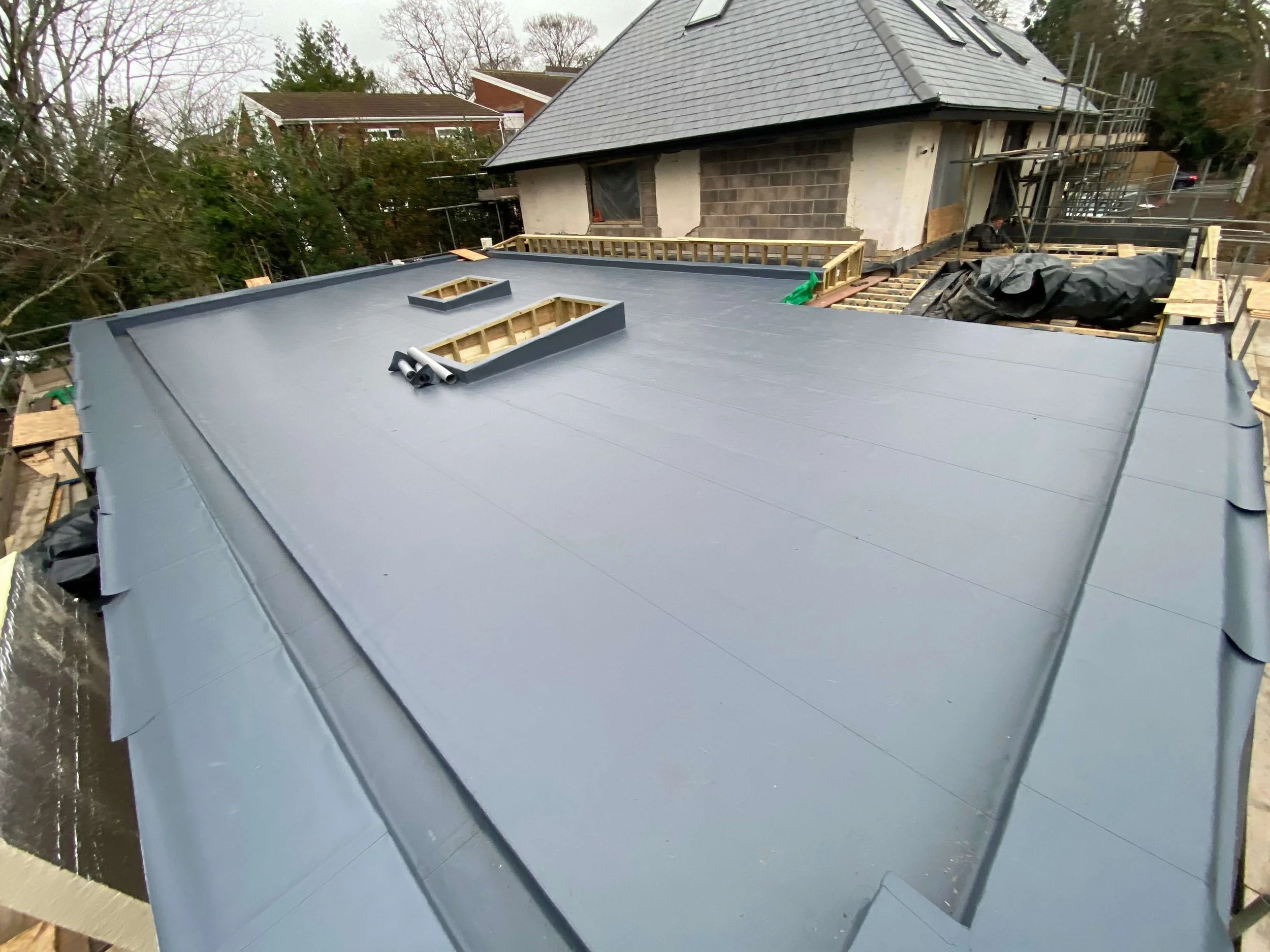 Construction workers installing a new flat roof on a house, with scaffolding and building materials visible.