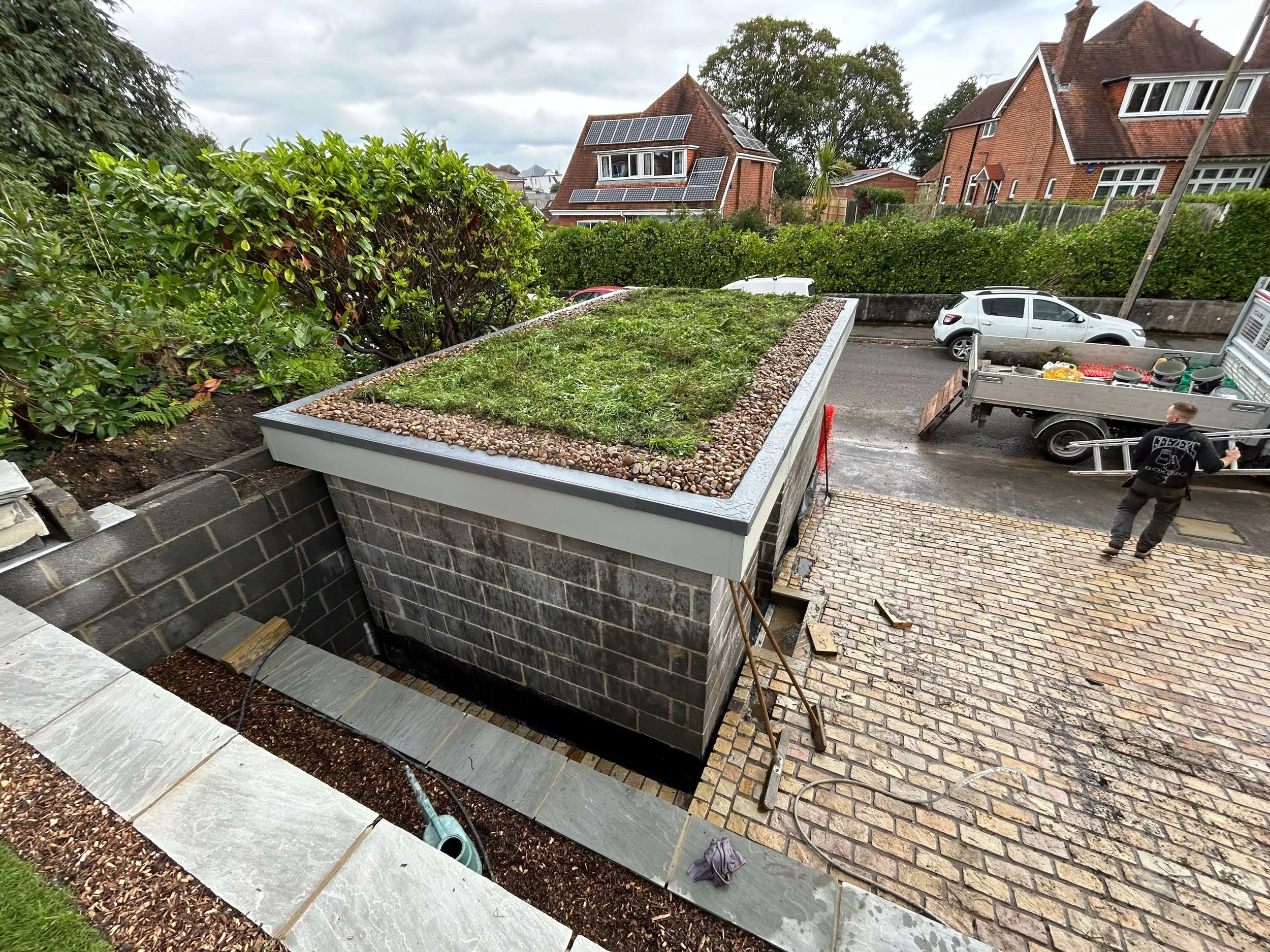View of a construction site in a residential neighborhood with a raised garden bed, construction tools, and workers. Houses with solar panels in the background.