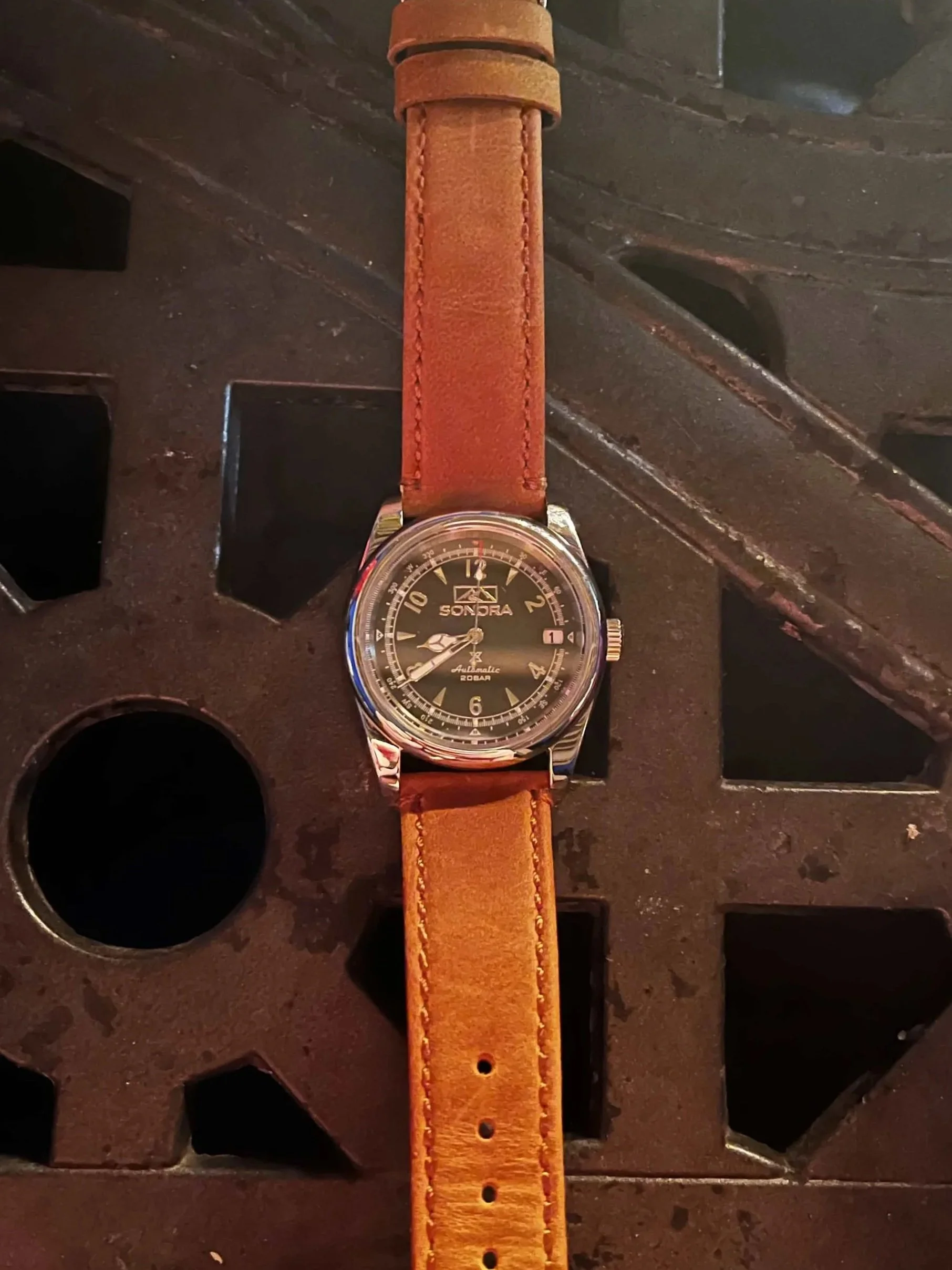 A silver oyster style seiko nh35 automatic wristwatch with a green vintage dial with a brown leather strap on a black metal surface with square cutouts.