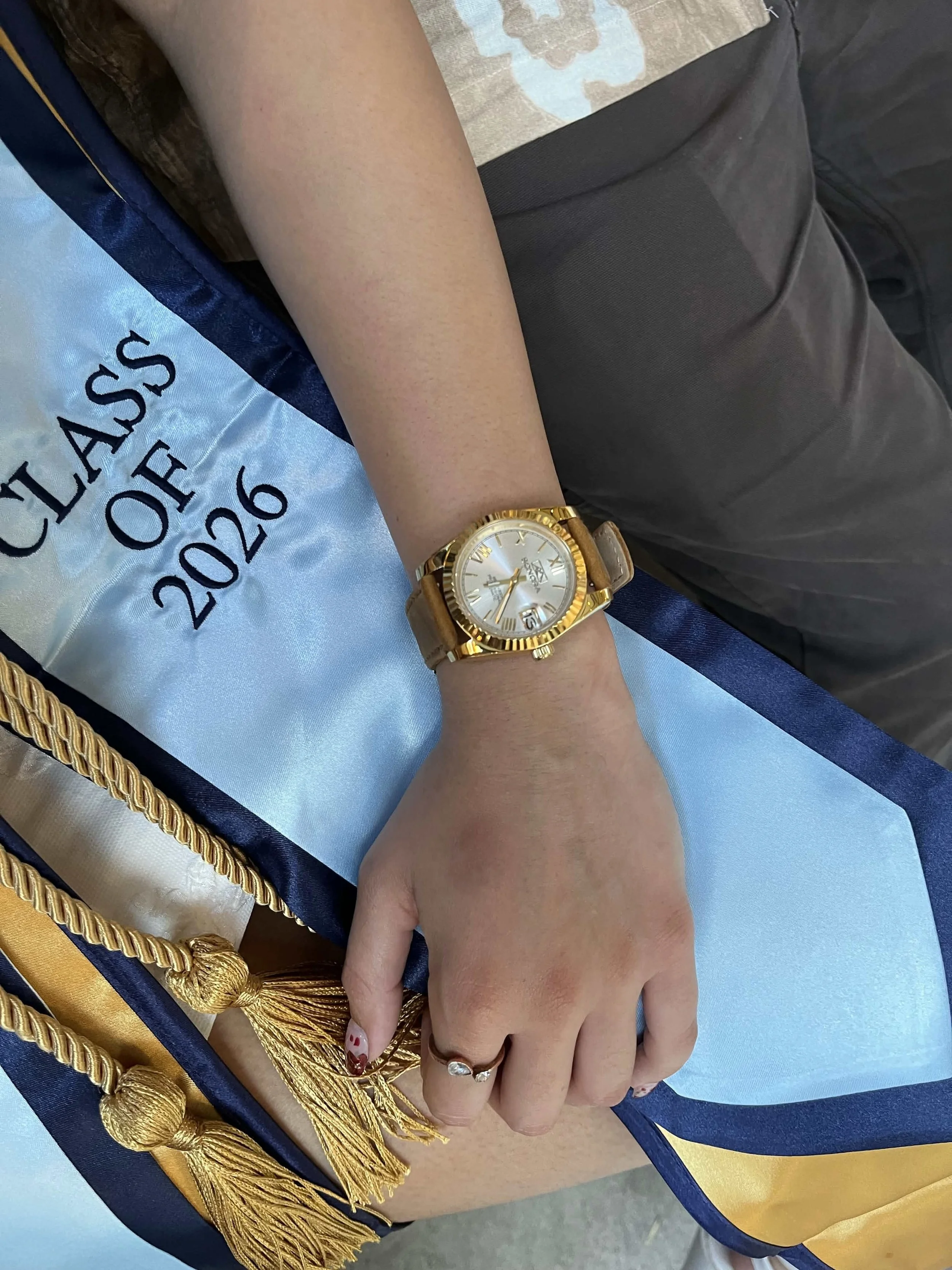 Close-up of a person's hand and wrist wearing an automatic seiko nh35 gold watch with a white face, showing Roman numerals. The person is wearing a ring. The watch is a graduation gift for a 2026 college graduate.