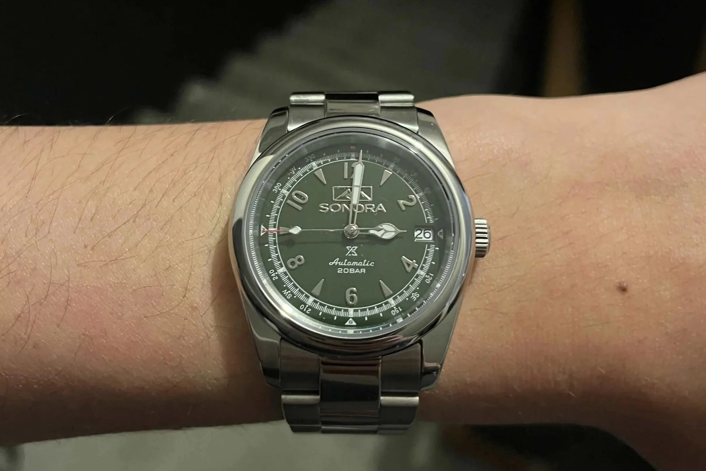 A person's wrist wearing a silver analog wristwatch with a black face, white markers, and a date window showing the 26th.