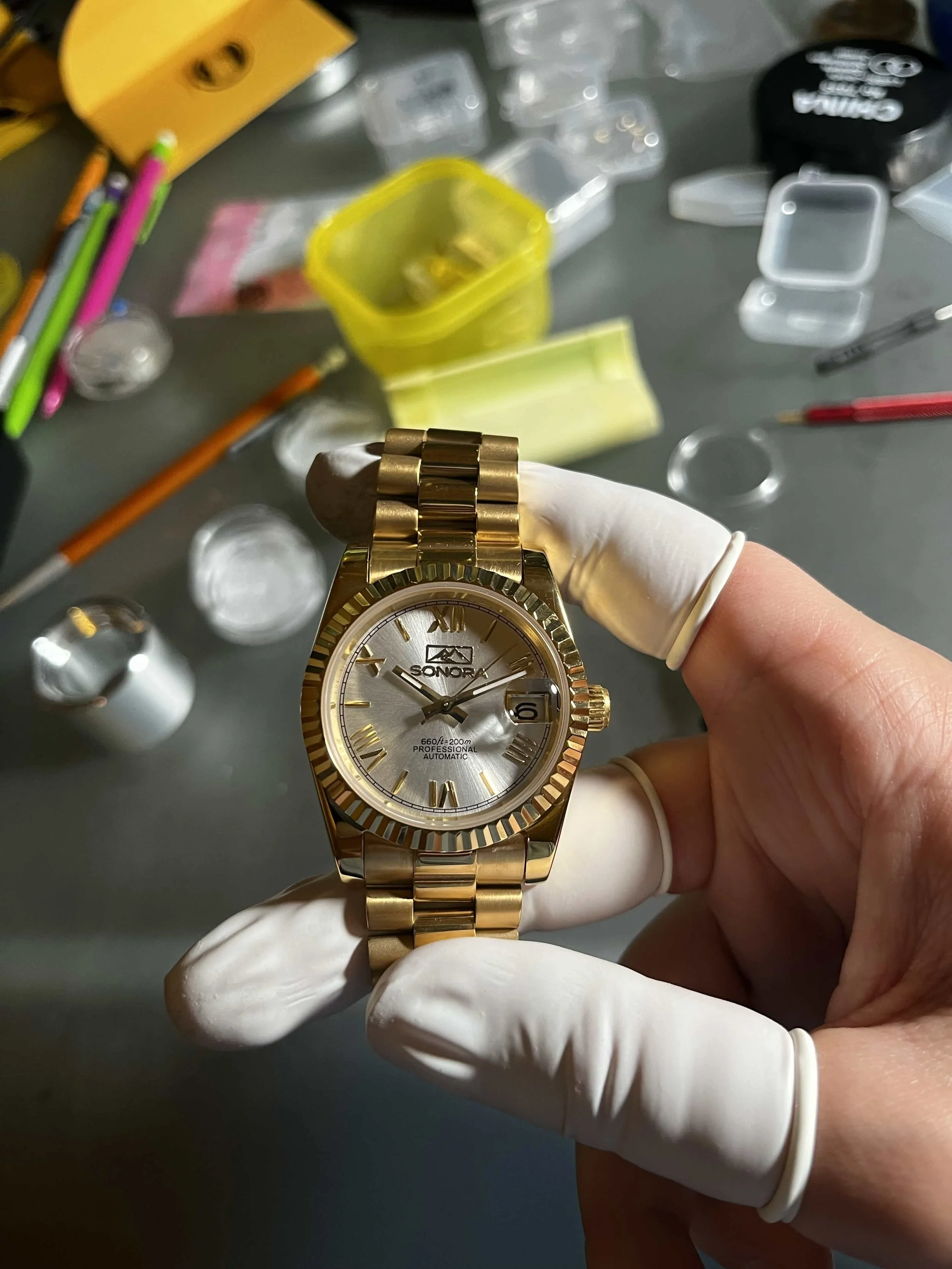 A gloved hand holding a gold-colored wristwatch with a silver dial that has Roman numerals and additional small dials, in a workspace with various small containers and tools scattered on a gray surface.