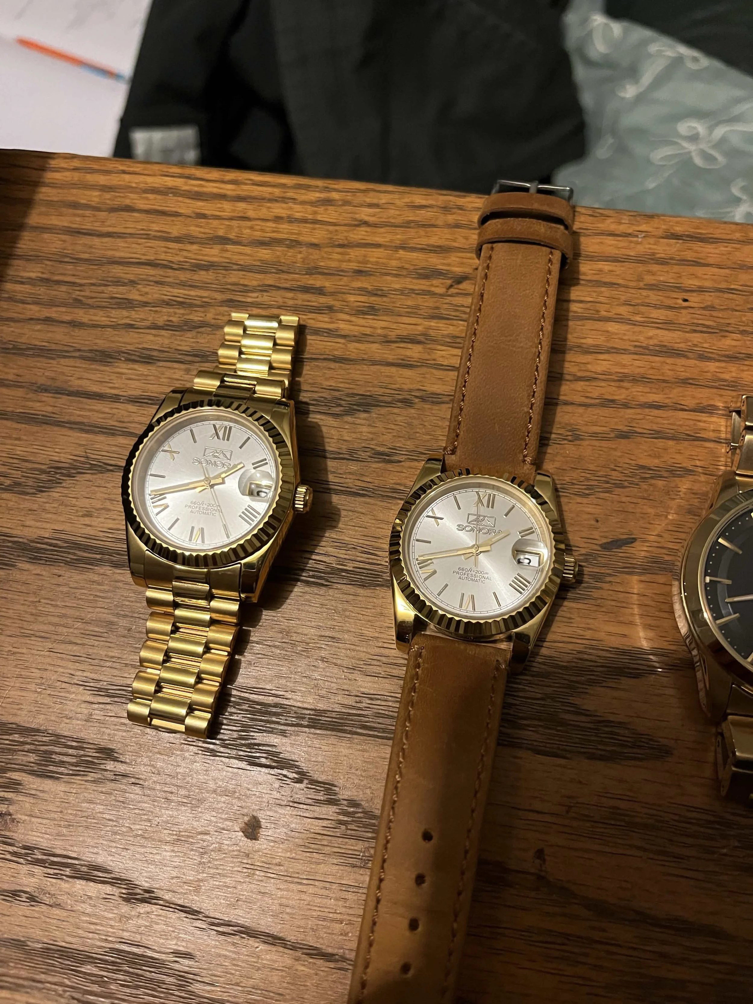 Two gold-colored wristwatches with white dials and Roman numerals, one with a gold metal band and the other with a brown leather strap, placed on a wooden surface.