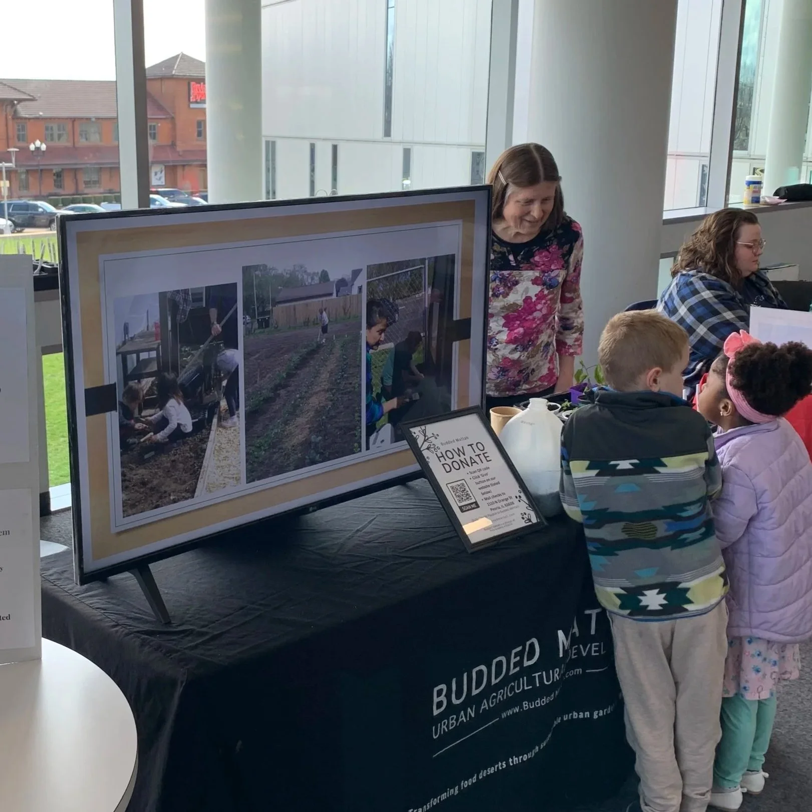 People at a table with a display board showing children working in a garden, and a sign with a QR code for donations, inside a building with large windows.