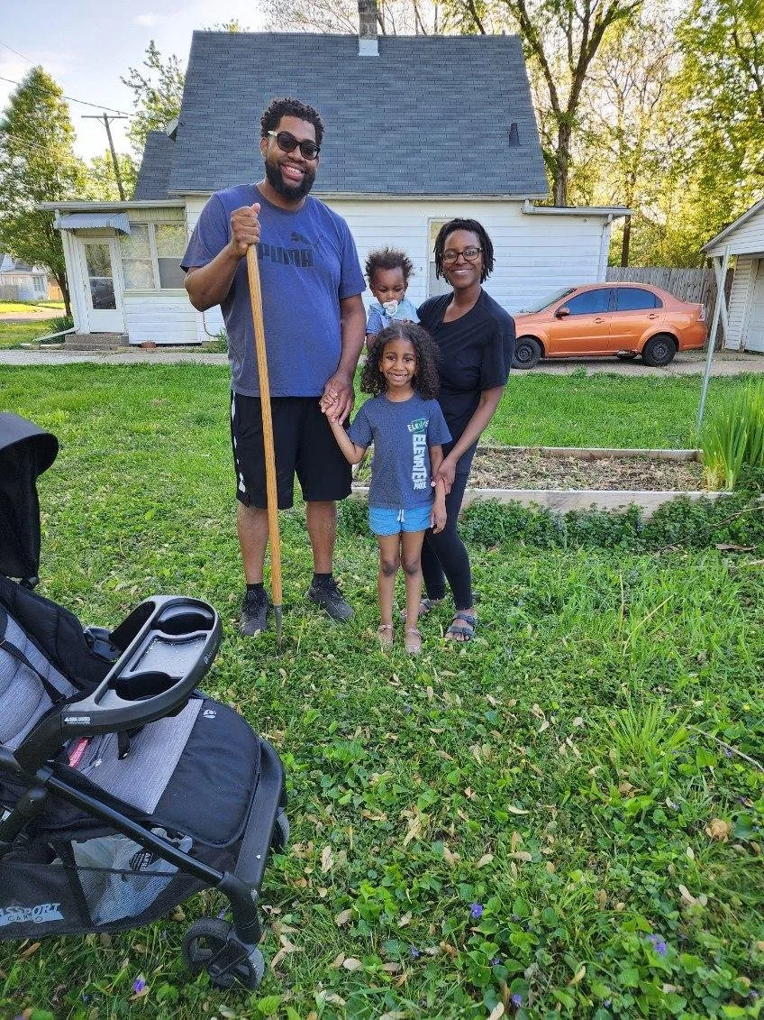 A family of four, including a man holding a gardening tool, a woman, and two young girls, posing in a backyard with a house, trees, and a parked car in the background.