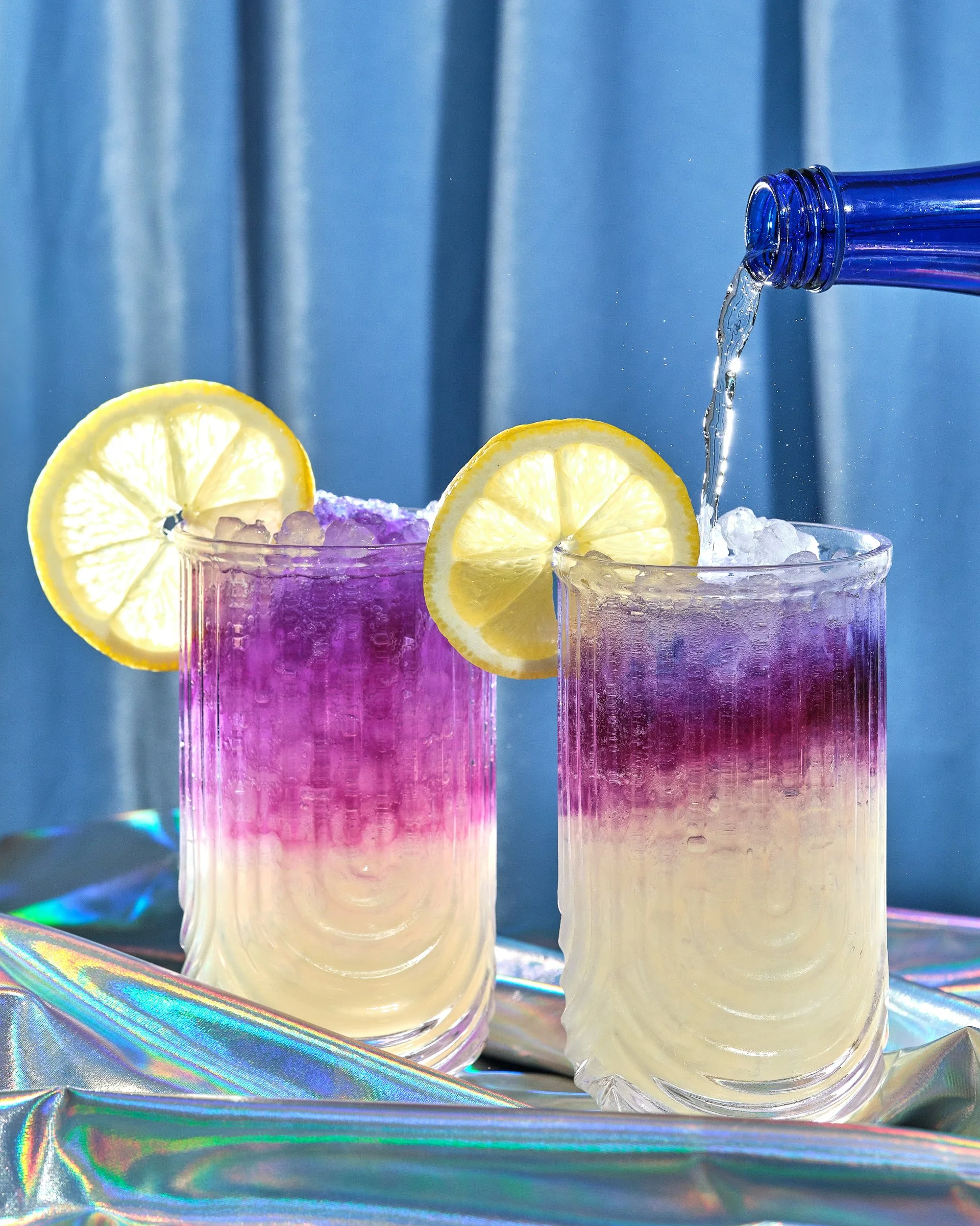 Two glasses of colorful beverage with lemon slices and ice, on a holographic surface with a blue background, with water pouring into the glass on the right.