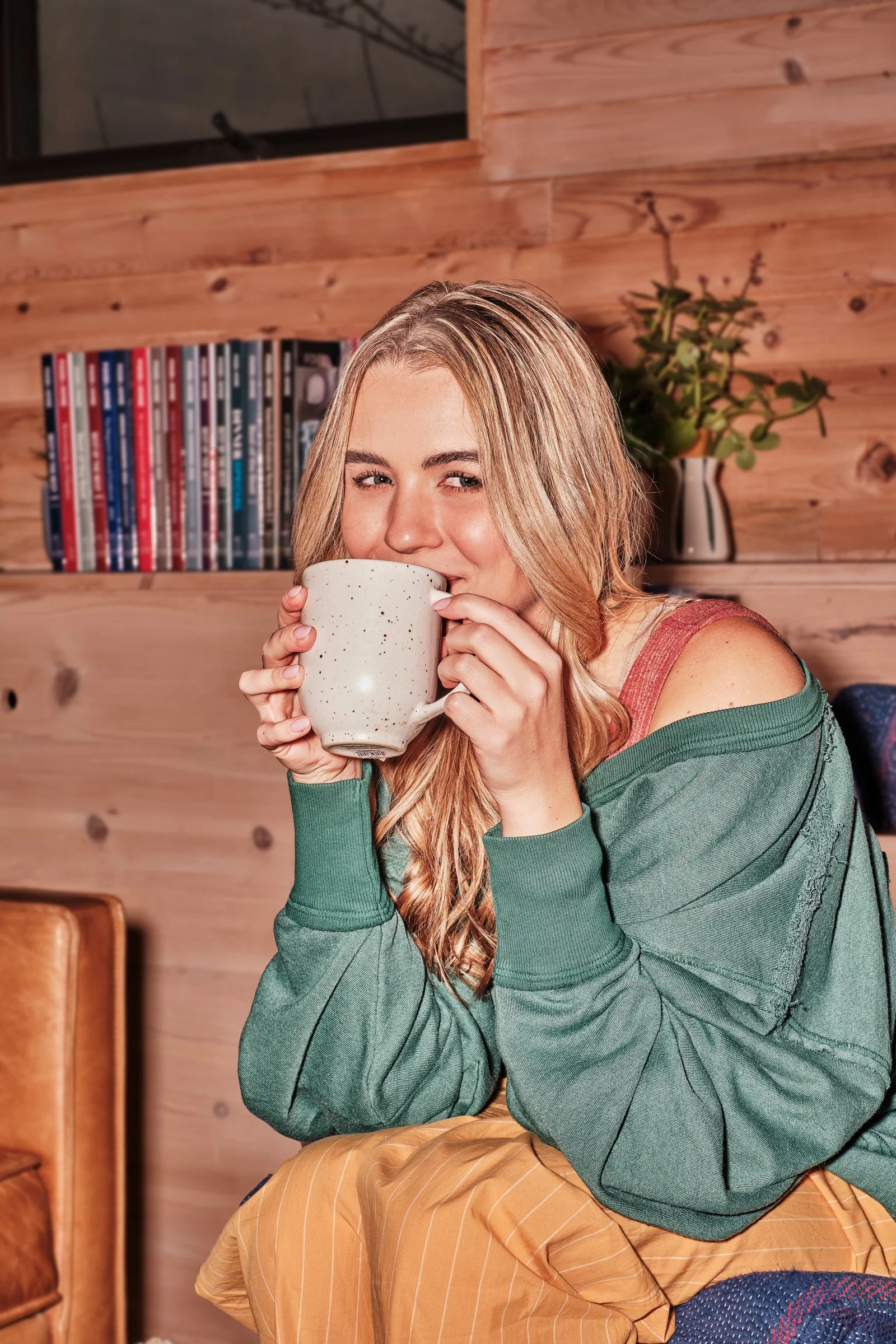 A young woman with long blonde hair, wearing a green sweater, is holding a white mug and smiling in front of a wooden wall with a bookshelf and a plant in a white vase.
