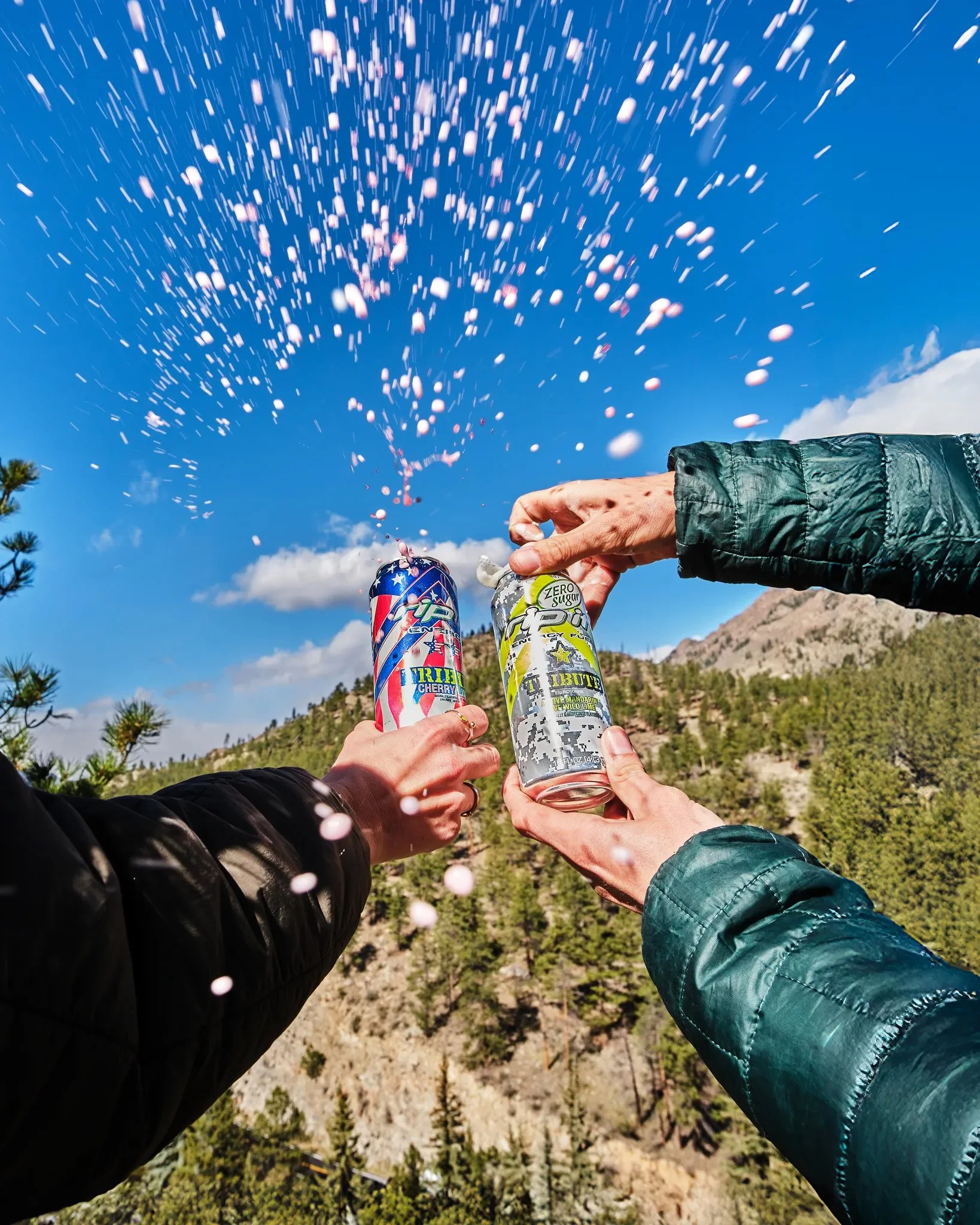 People holding cans of energy drinks and champagne bottles during daytime in an outdoor setting with mountainous terrain and a partly cloudy sky.