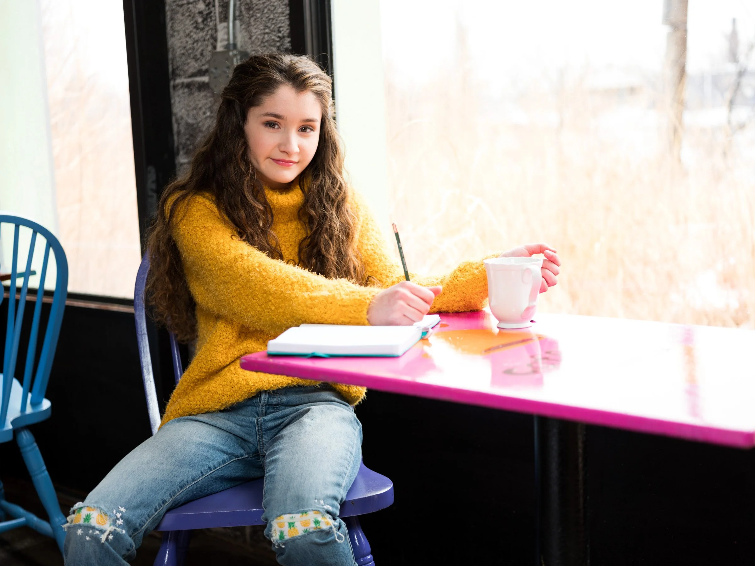 Young girl with long curly hair wearing a yellow sweater and jeans, sitting at a colorful table with a pink surface, holding a paintbrush near a white ceramic mug, with a notebook and colored paper on the table, indoors near a large window.