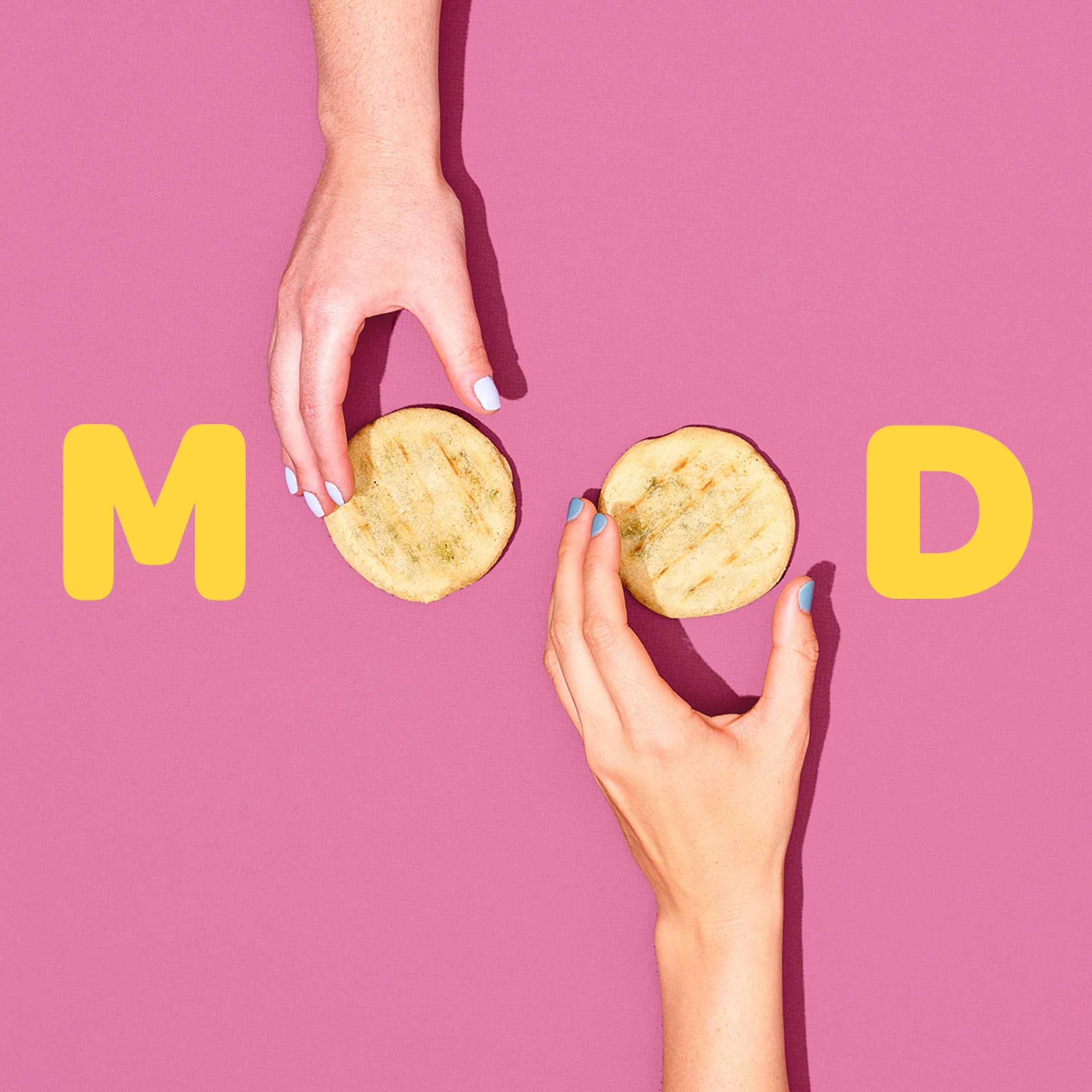 A hand with manicured nails is holding a golden brown cookie on a pink background, with a portion of yellow text visible at the bottom.