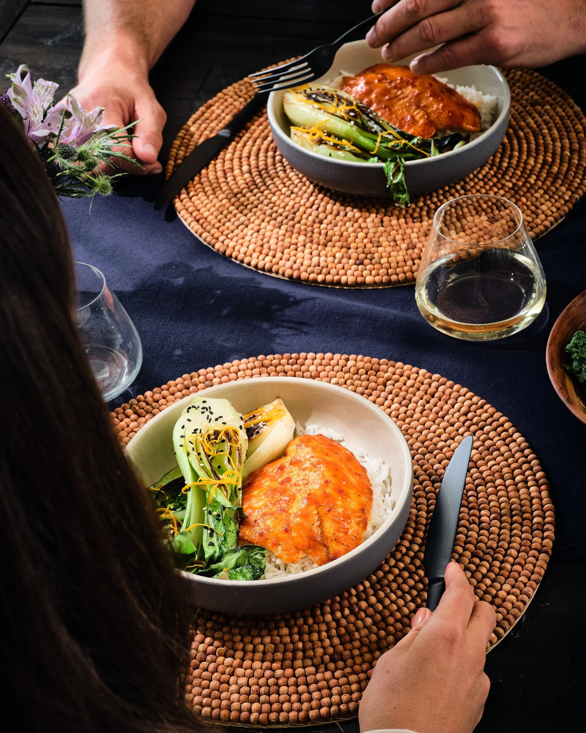 Person holding a fork and knife, serving a bowl of white rice topped with a piece of glazed salmon, grilled green vegetables, and garnishes, on a table with a woven placemat, wine glass, and floral centerpiece.