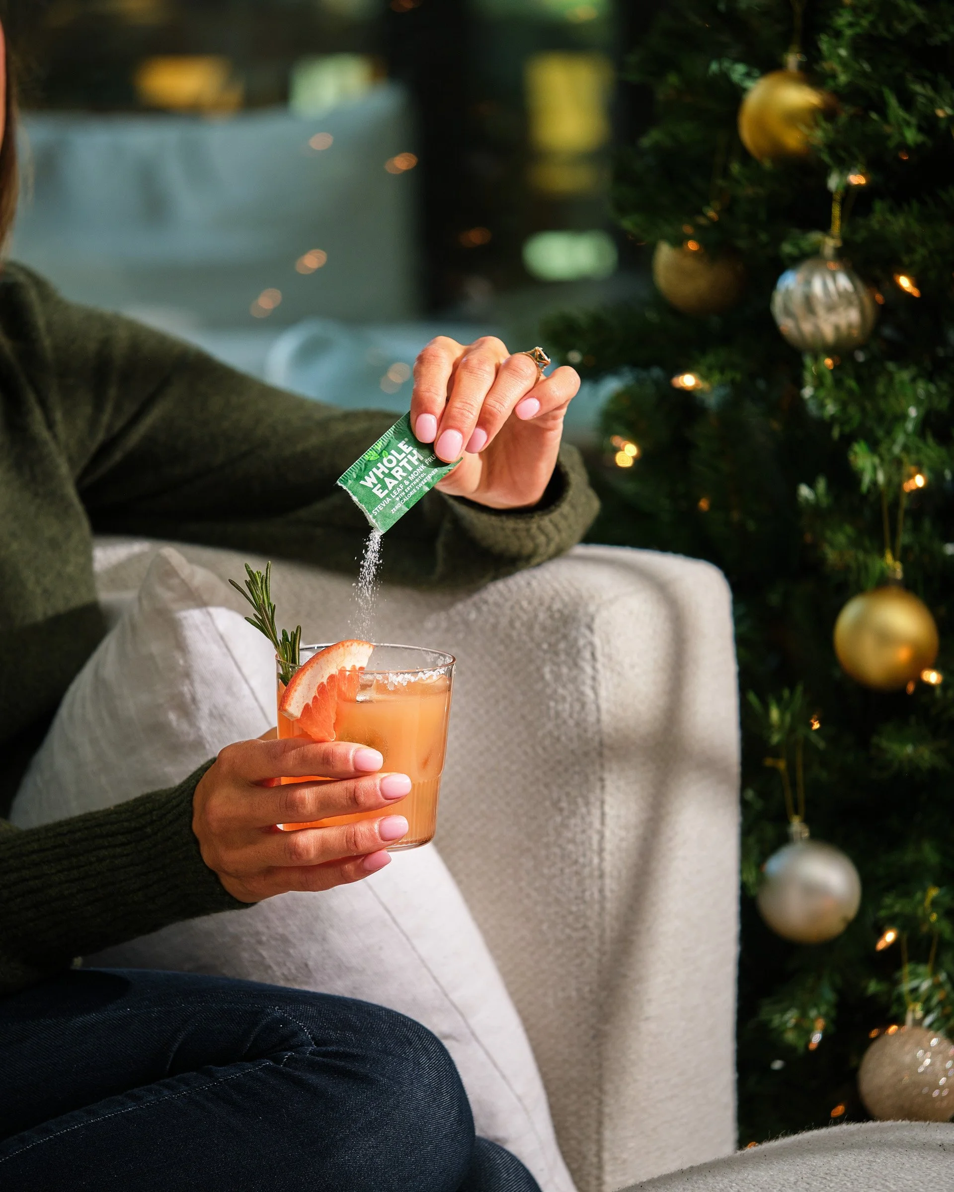 Person holding a glass of orange-colored beverage garnished with a strawberry, rosemary sprig, and a slice of orange, while sprinkling a green packet labeled 'Whole Earth Stevia Leaf' powder into it. A Christmas tree with gold and silver ornaments is
