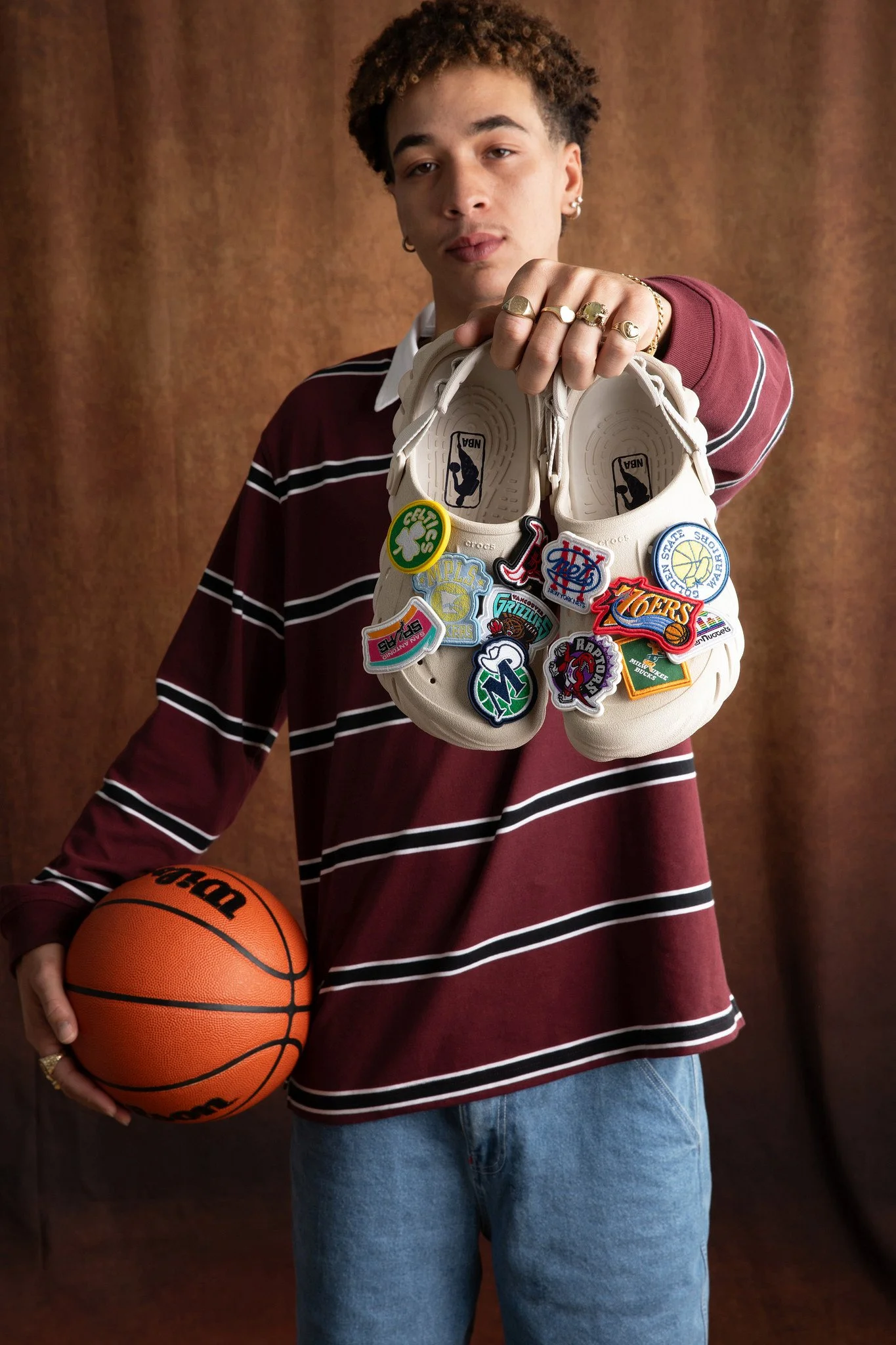 A person wearing a maroon and white striped shirt holding an orange basketball in one hand and a pair of white sneakers with NBA patches in the other hand, standing in front of a brown backdrop.